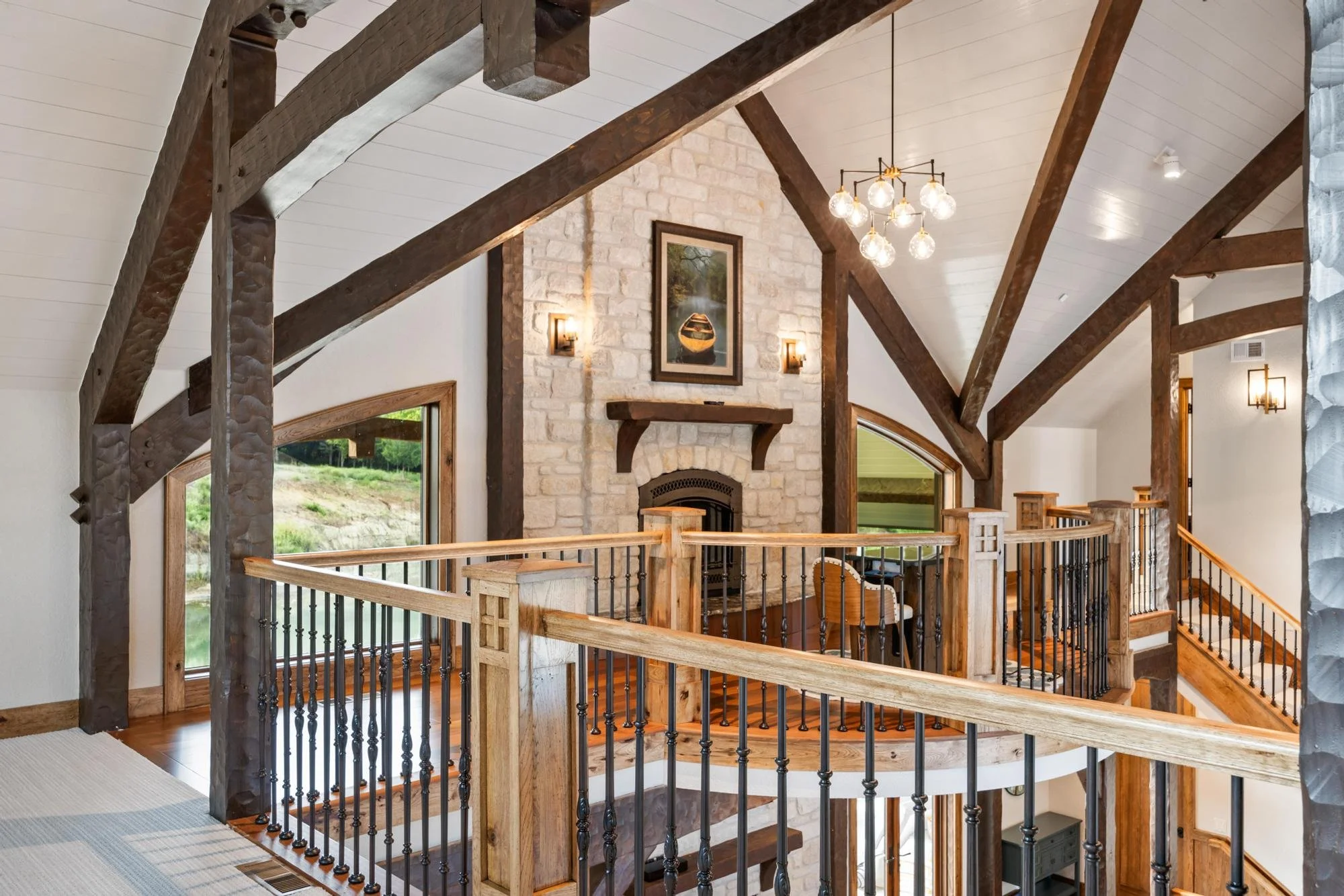 Interior view of a rustic home with exposed wooden beams, stone fireplace, and large windows, featuring a chandelier hanging from the vaulted ceiling.