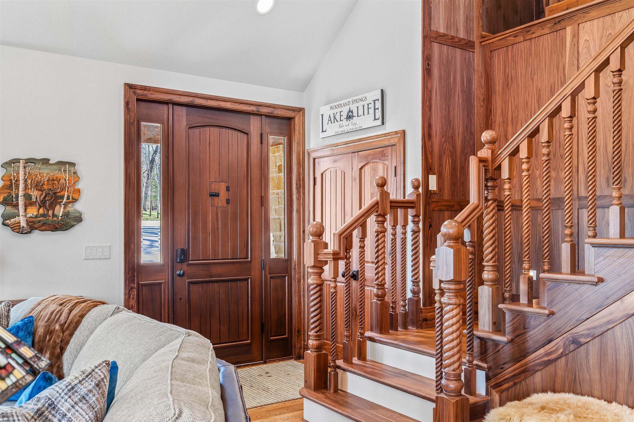 Interior of a house showing a wooden front door with glass panels, a staircase with turned wooden balusters, a decorative wall art of a moose and trees, and a white wall with a sign that reads 'Woodland Springs Lake Life'.