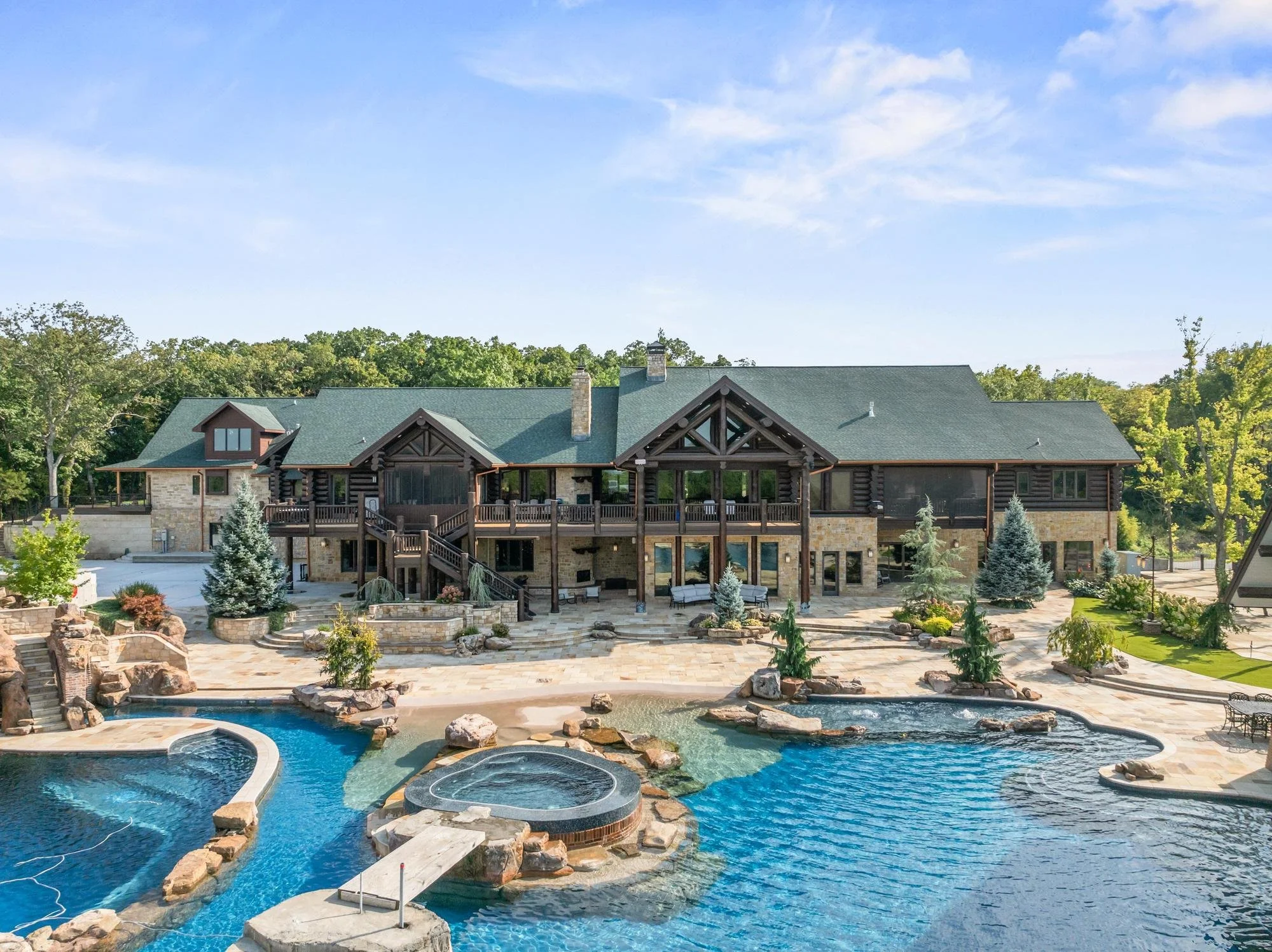 Large house with green roofs, stone and dark wood exterior, surrounded by trees, with a swimming pool and hot tub in the foreground.