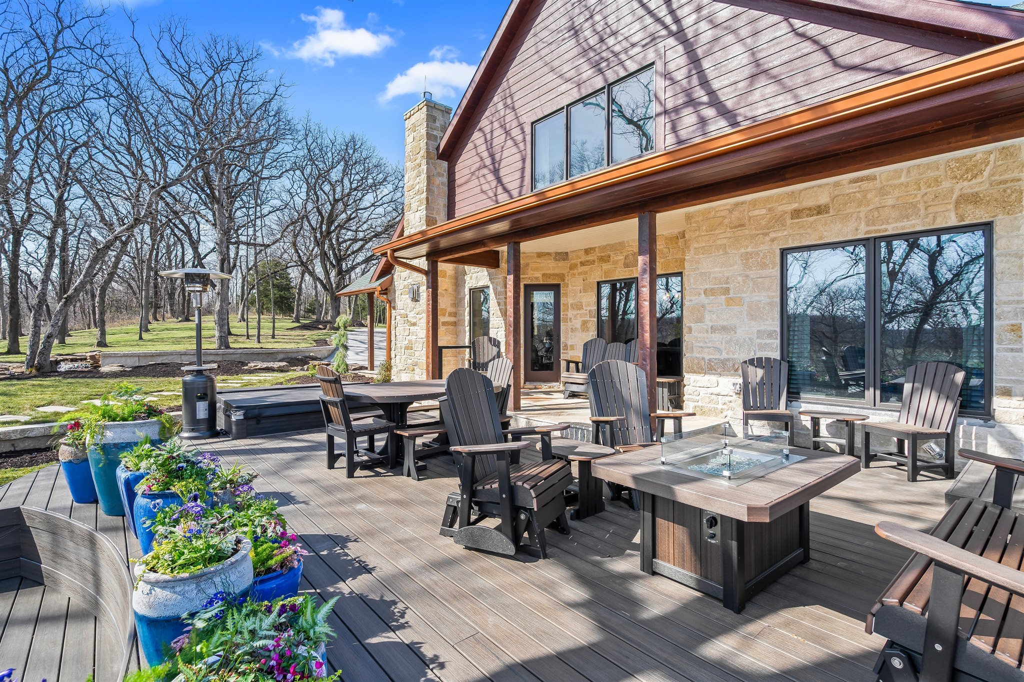 Outdoor patio area with wooden decking, black Adirondack chairs, a glass table, and colorful potted flowers. House exterior features stone and wood siding, large windows, and a chimney. Trees in the background under a blue sky.