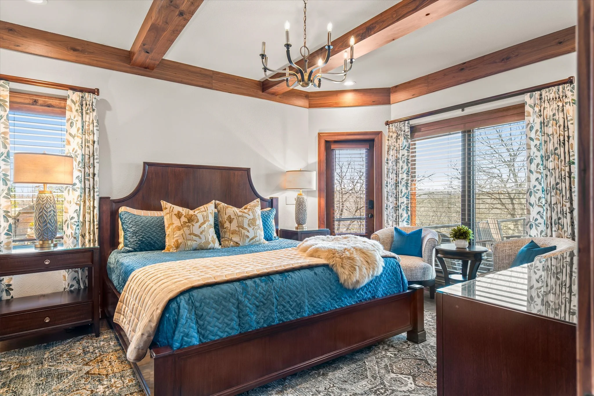 Bedroom with wooden bed frame, blue and beige bedding, and decorative pillows, large windows with floral curtains, wooden ceiling beams, chandelier, side table with lamp, and sitting area with small table and chairs.