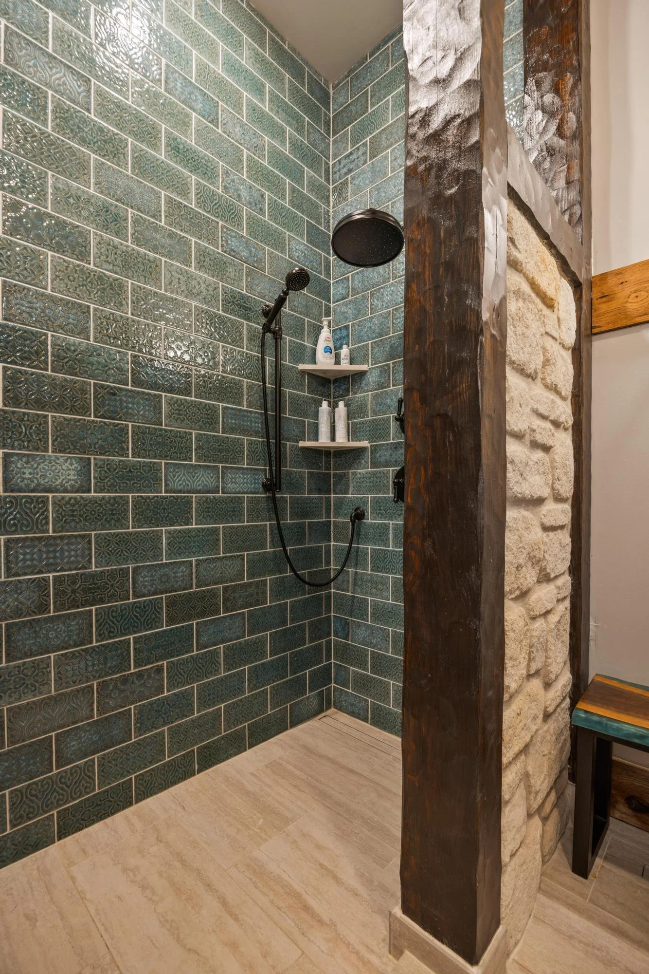 A bathroom shower area with dark green patterned tiles, a black showerhead, a small white tile shelf holding toiletries, a rustic wooden and stone wall, a small colorful bench, and beige tile flooring.