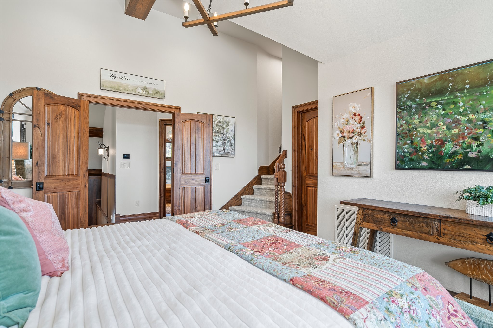 A bedroom with white walls, wooden trim, and artwork. There is a bed with a white quilt, pink and teal pillows, and a quilted bed runner. The room features wooden accents, a staircase, and colorful paintings on the wall.