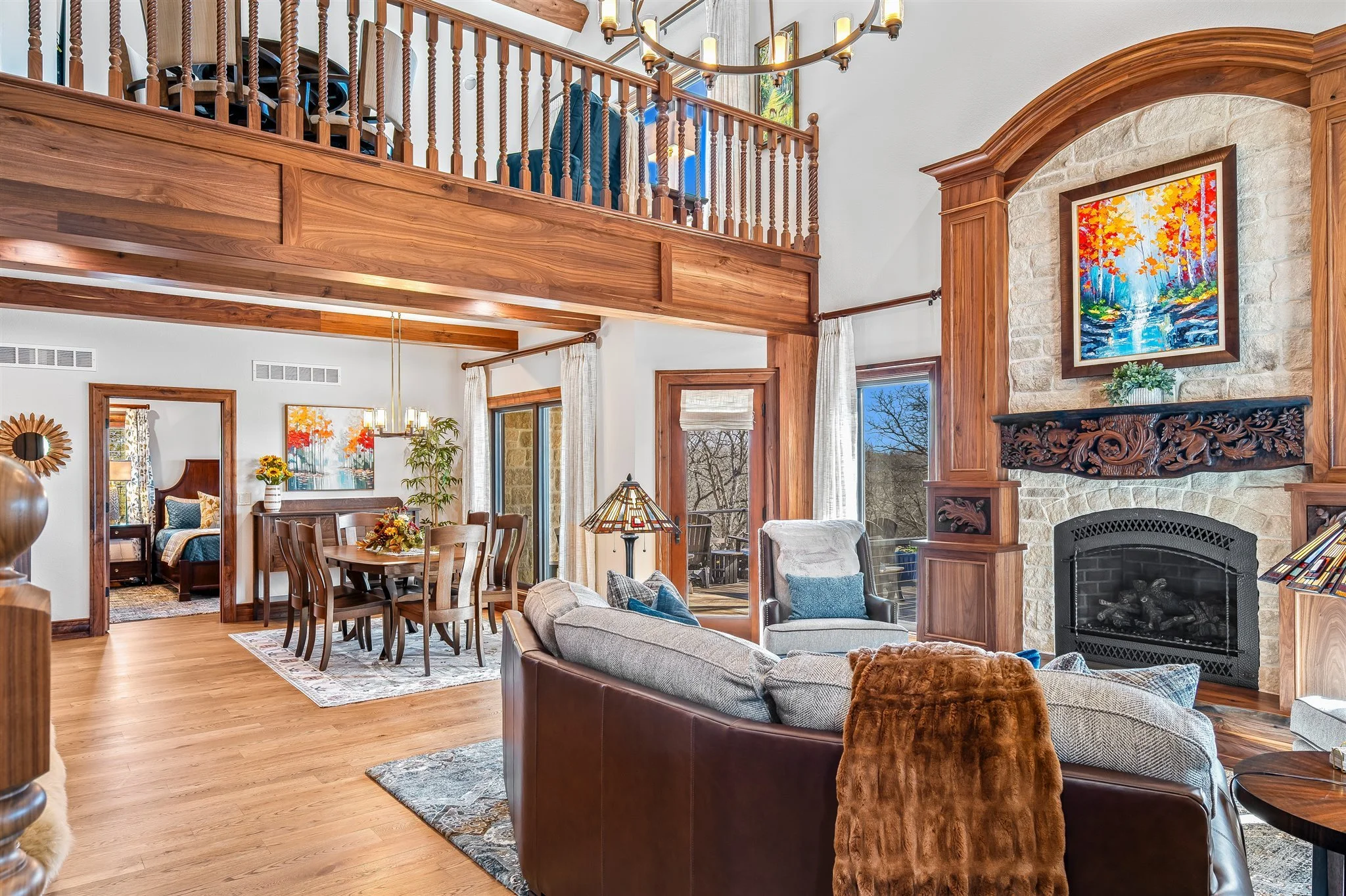 A spacious living room with a high vaulted ceiling, large windows, and a loft area with wooden railing. There is a stone fireplace with ornate woodwork and a colorful painting above it. The room has hardwood floors, a sofa, armchairs, and a dining ta