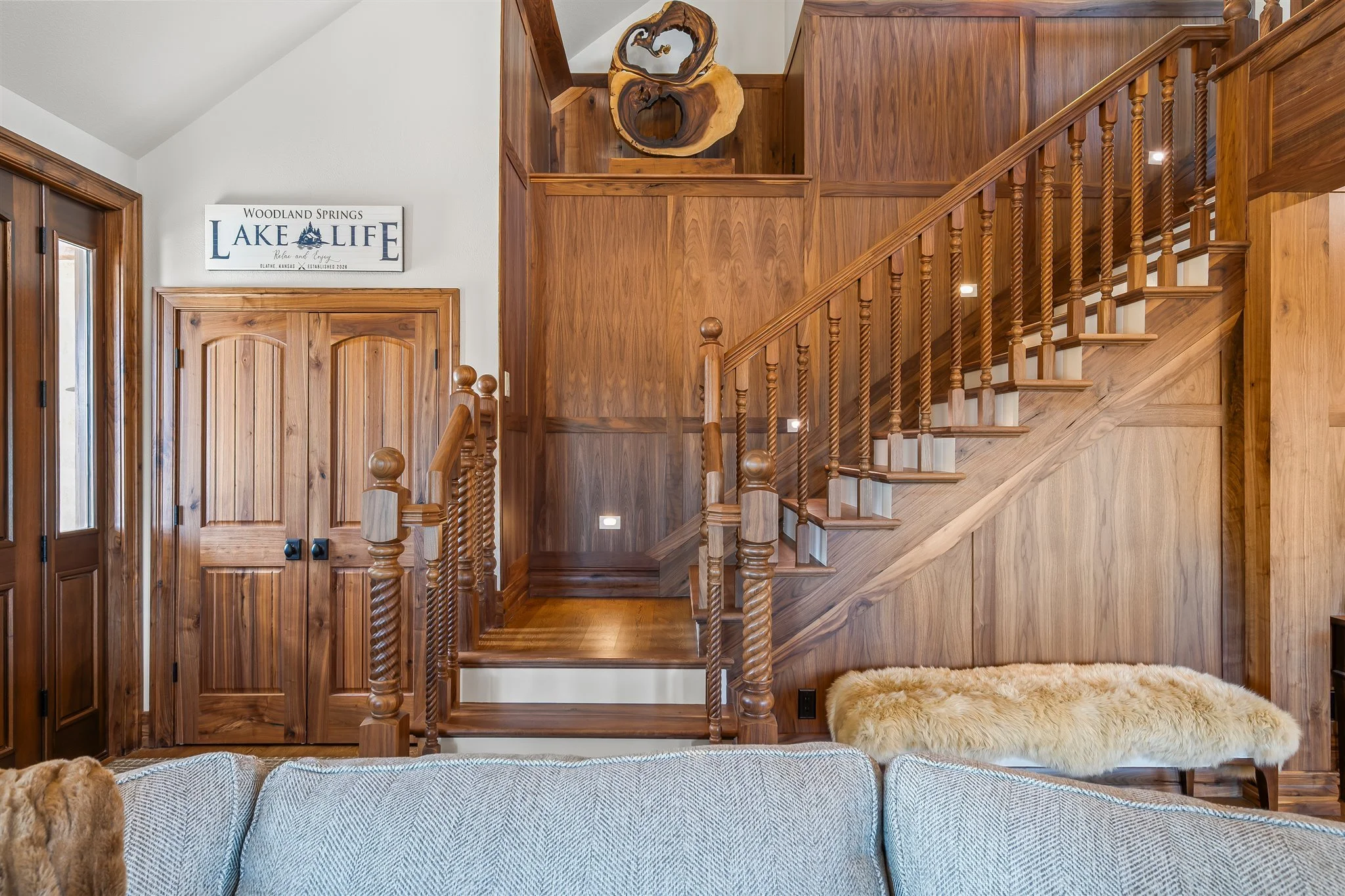 Interior view of a wooden staircase with a wooden railing, a cabinet with wooden doors, a sign that says 'Woodland Springs Lake Life,' and a cozy beige rug.