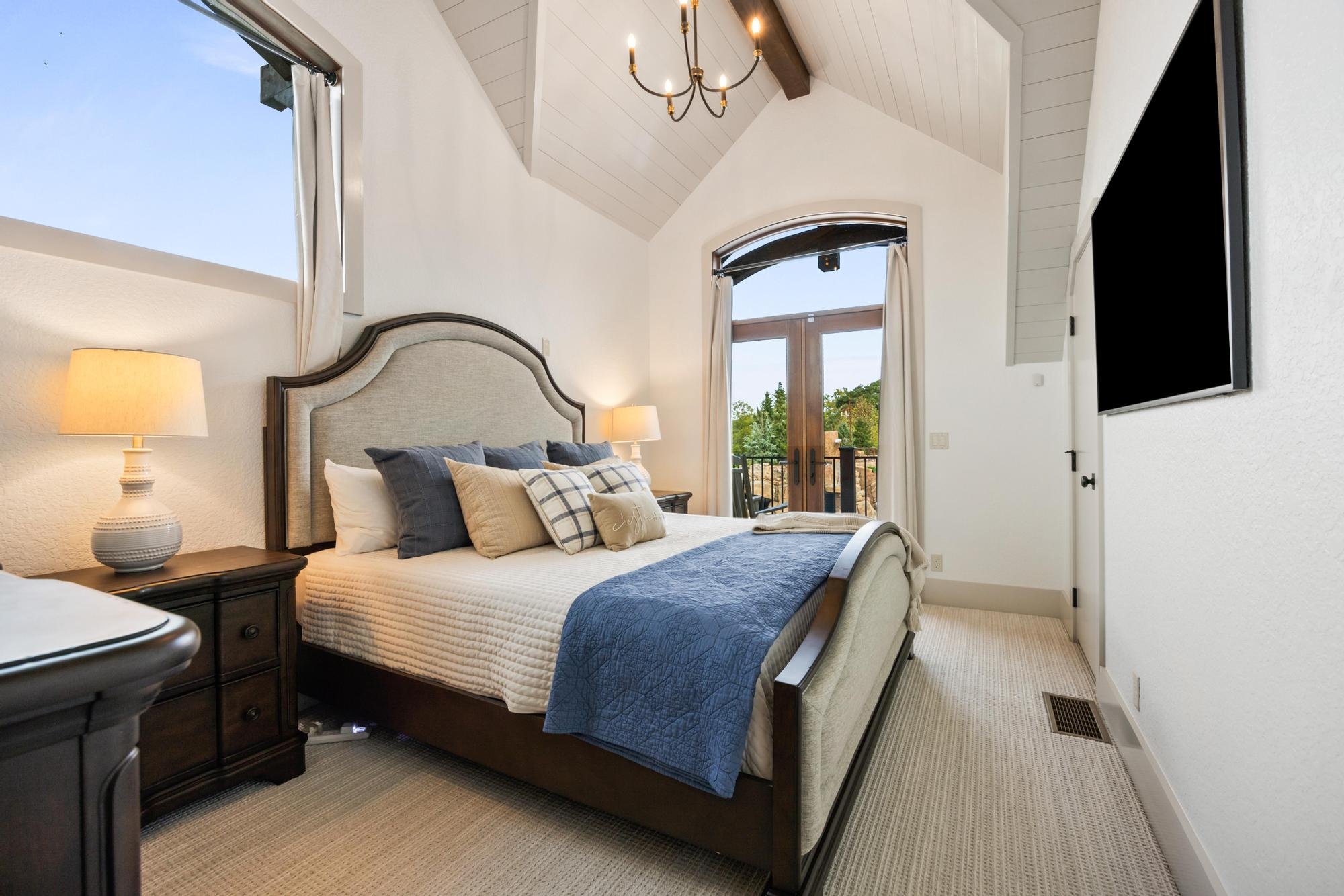 A bedroom with a vaulted ceiling, chandelier, large window with curtains, and a door leading outside. The bed has multiple pillows and a blue quilt, and there is a nightstand with a lamp.