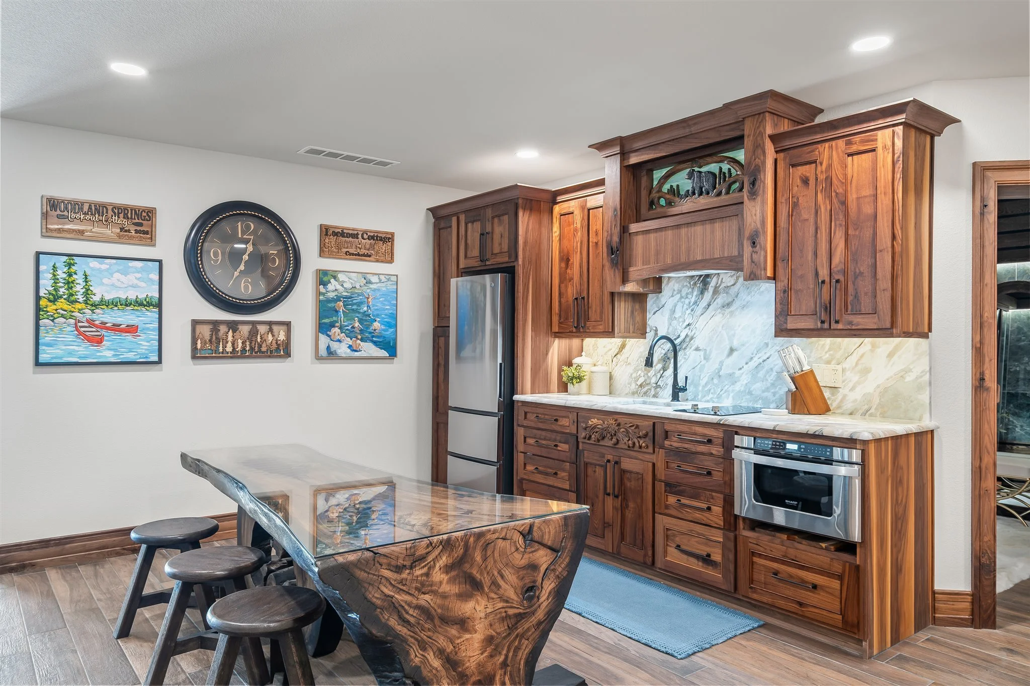 A kitchen with wooden cabinets, a marble backsplash, and a refrigerator. Wall art featuring outdoor scenes and a large clock, with a wooden dining table and three stools.