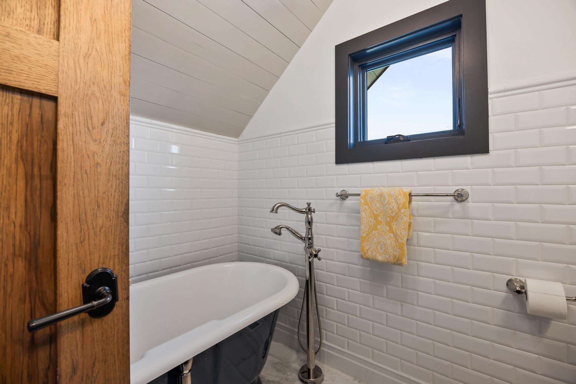 Bathroom with white subway tile walls, small bathtub, window with dark frame, yellow patterned towel hanging on a towel bar, and a silver faucet.