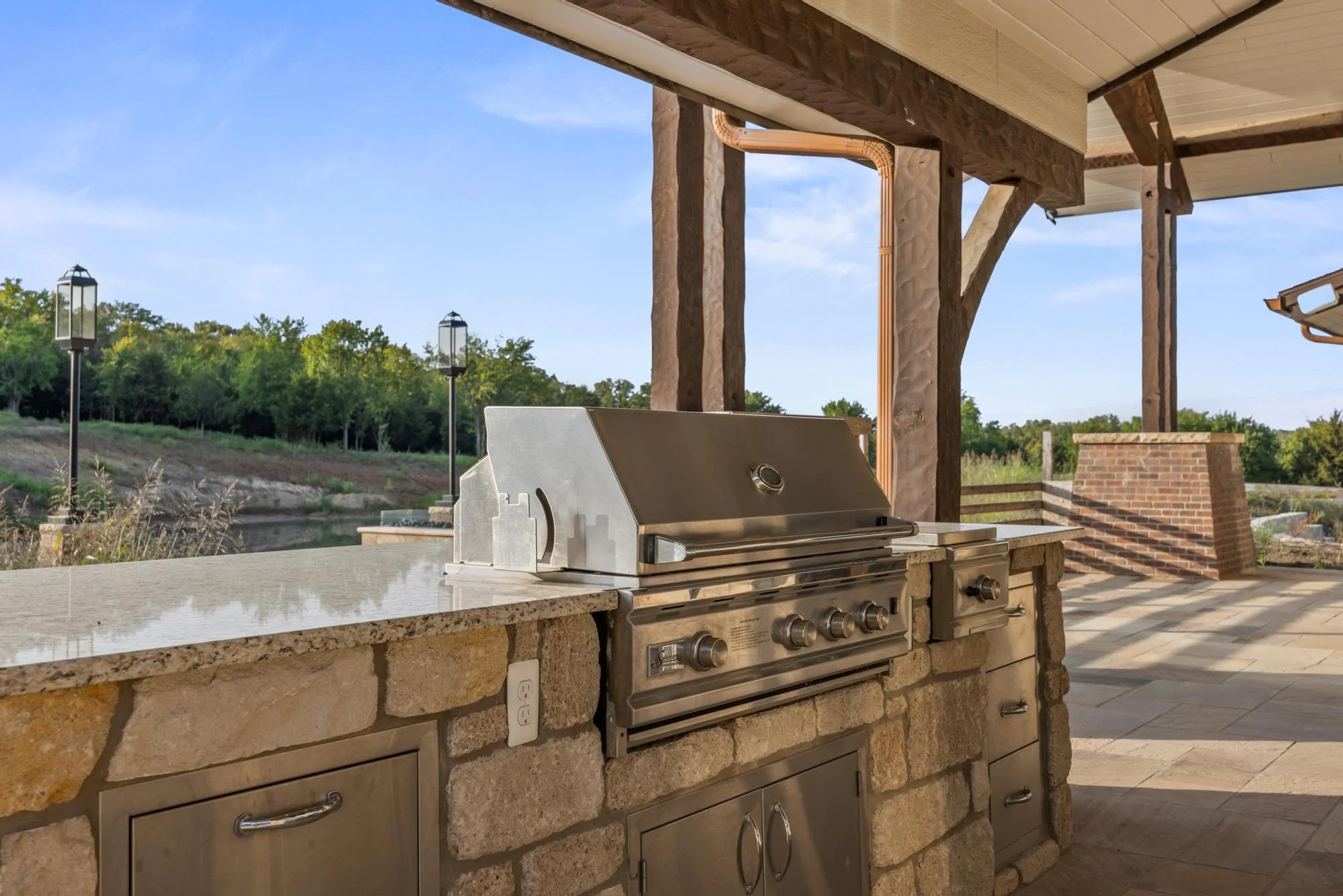 An outdoor kitchen with a built-in grill, stone countertop, and overhead wooden pergola. There are trees and a clear blue sky in the background. The kitchen features a stainless steel grill, brick accents, and decorative street lamps nearby.