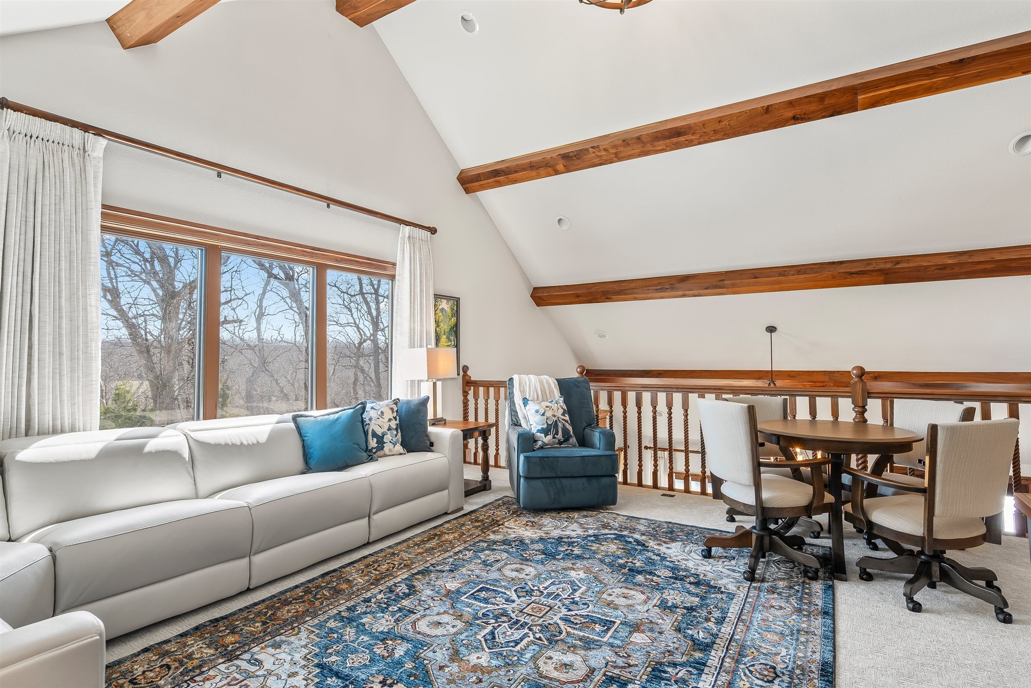 Living room with large window, white couch with blue and patterned pillows, blue armchair, area rug, ceiling beams, and a dining table with chairs.