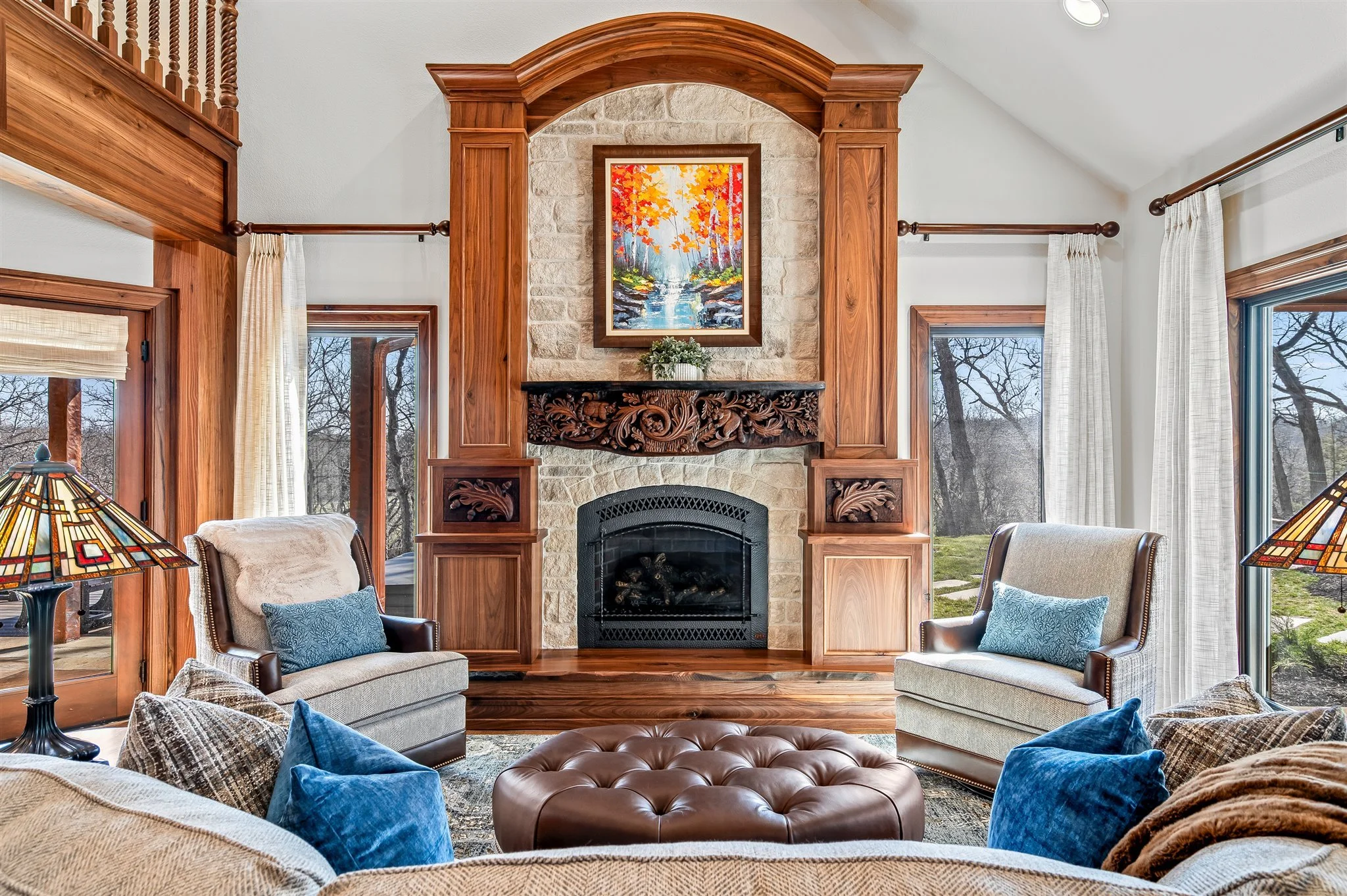 Living room with a stone fireplace, wooden mantel, and colorful painting above, surrounded by armchairs with pillows, large windows with curtains, and trees outside.