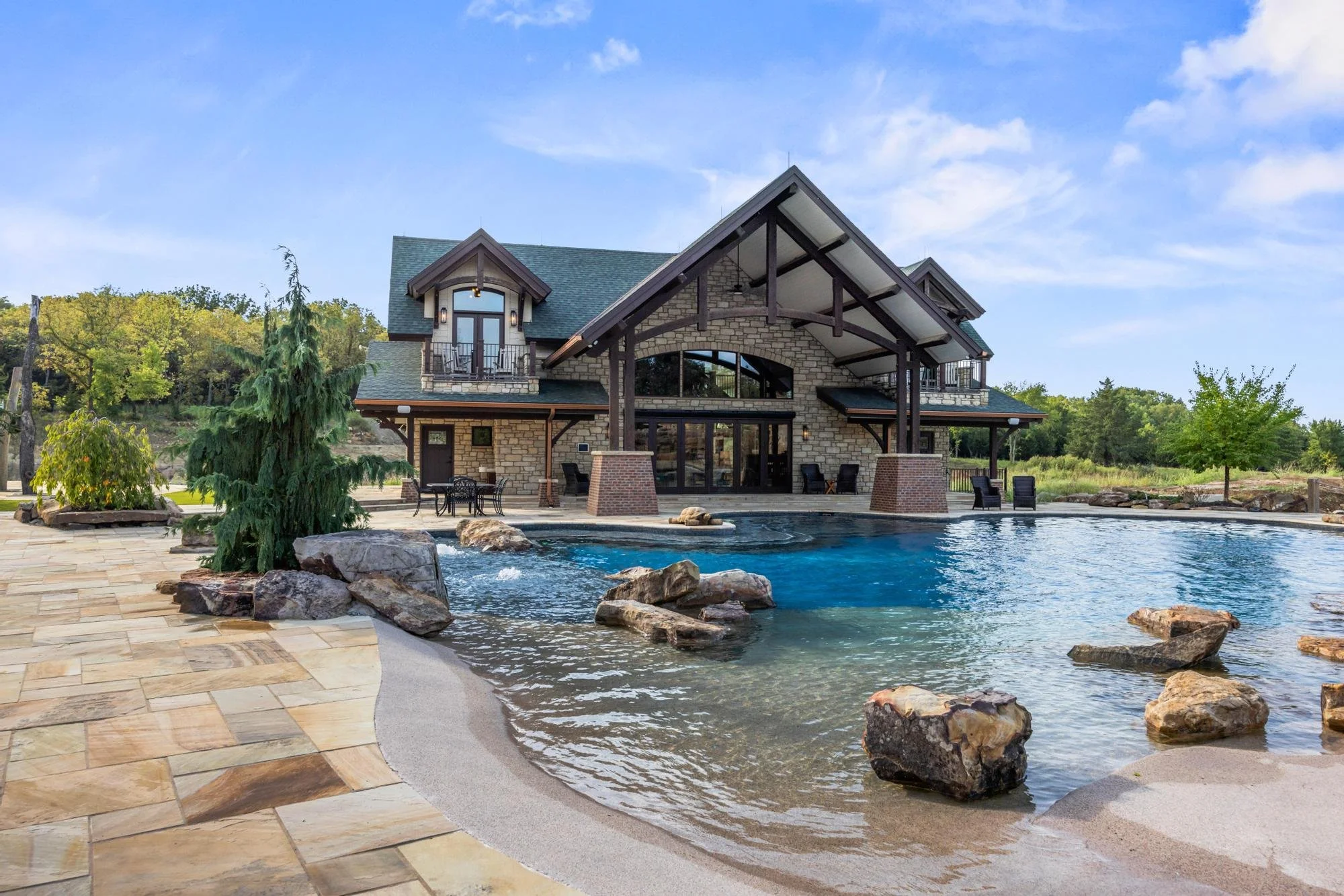 A large house with a stone and wood exterior and multiple balconies, situated by a swimming pool with rocks and trees around, under a partly cloudy sky.