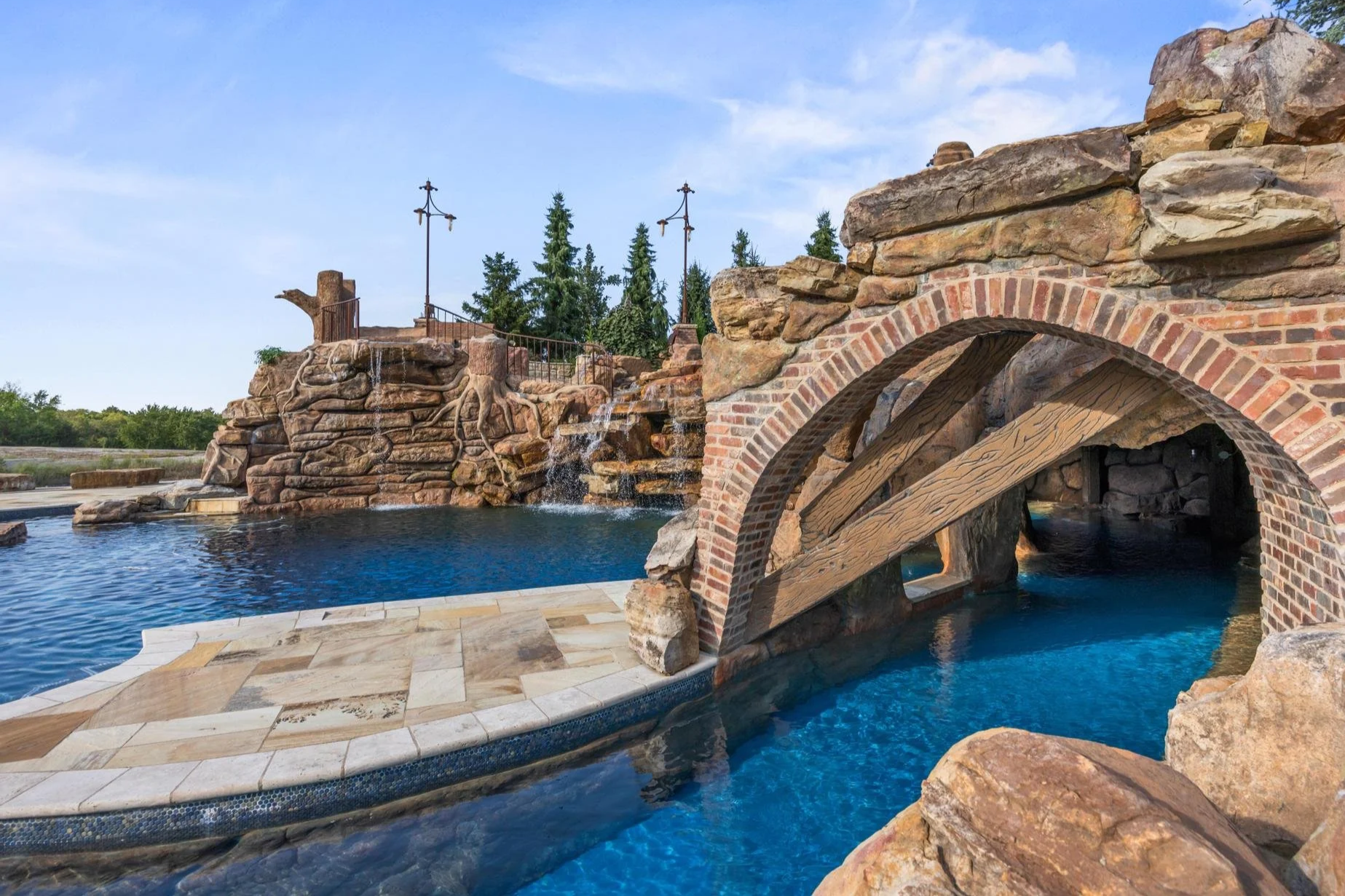 A scenic outdoor swimming pool area featuring a stone and brick bridge over the pool, with rock formations and a small waterfall in the background, surrounded by trees and a blue sky.