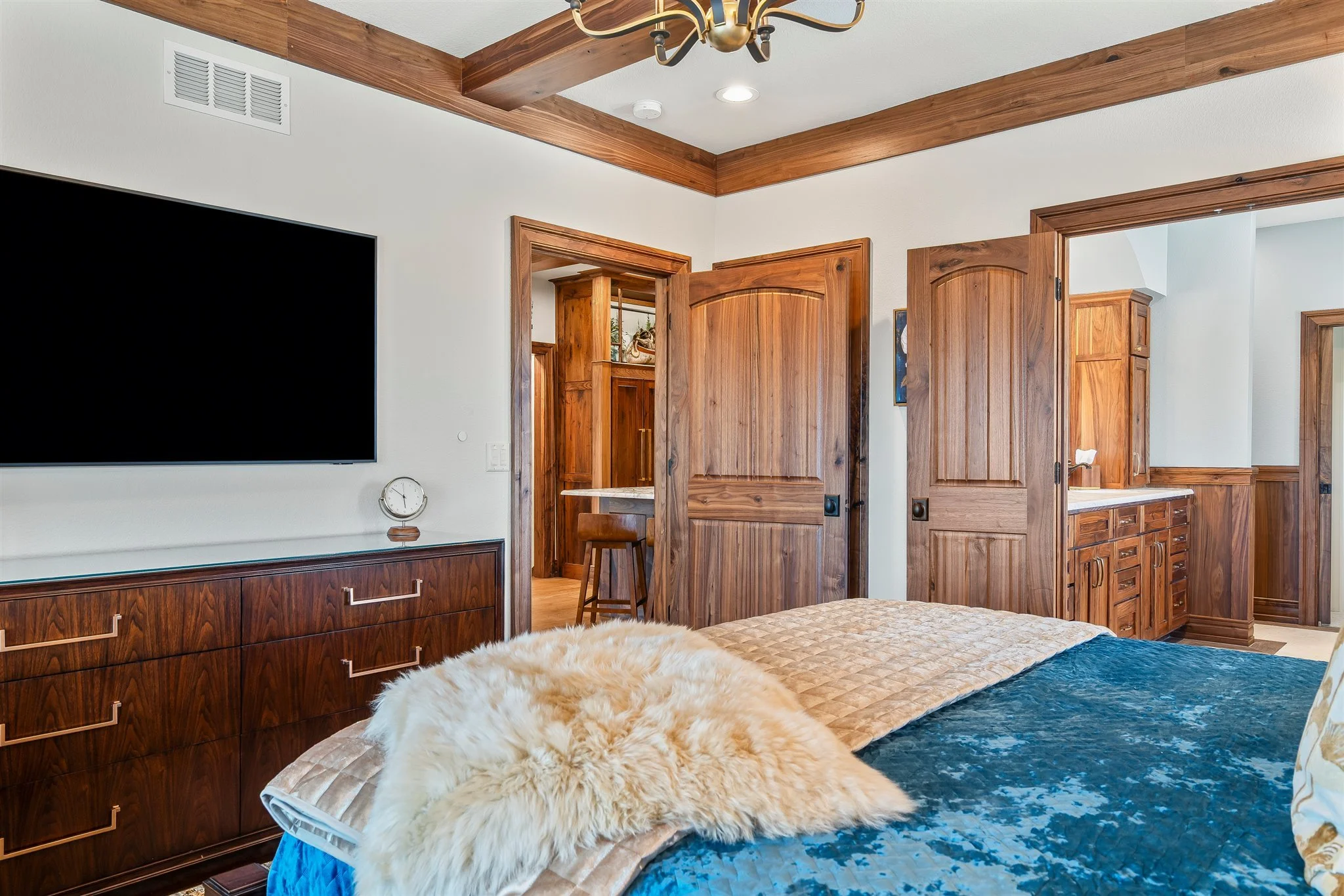 A bedroom with wood trim and cabinetry, a large flat-screen TV on the wall, a fur throw on the bed, and a chandelier overhead.