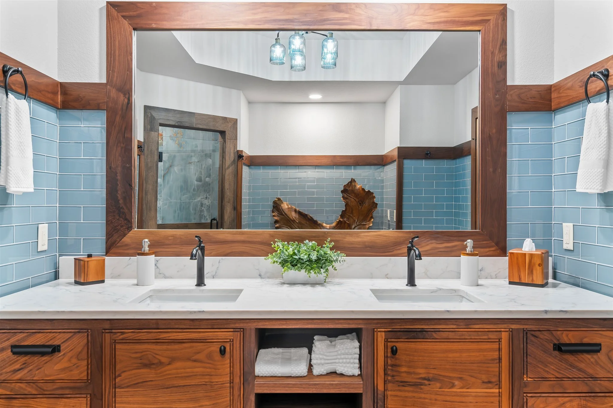 Bathroom vanity with a large mirror, wooden frame, a white marble countertop, black fixtures, a plant, soap dispensers, and light blue subway tiles on the walls.