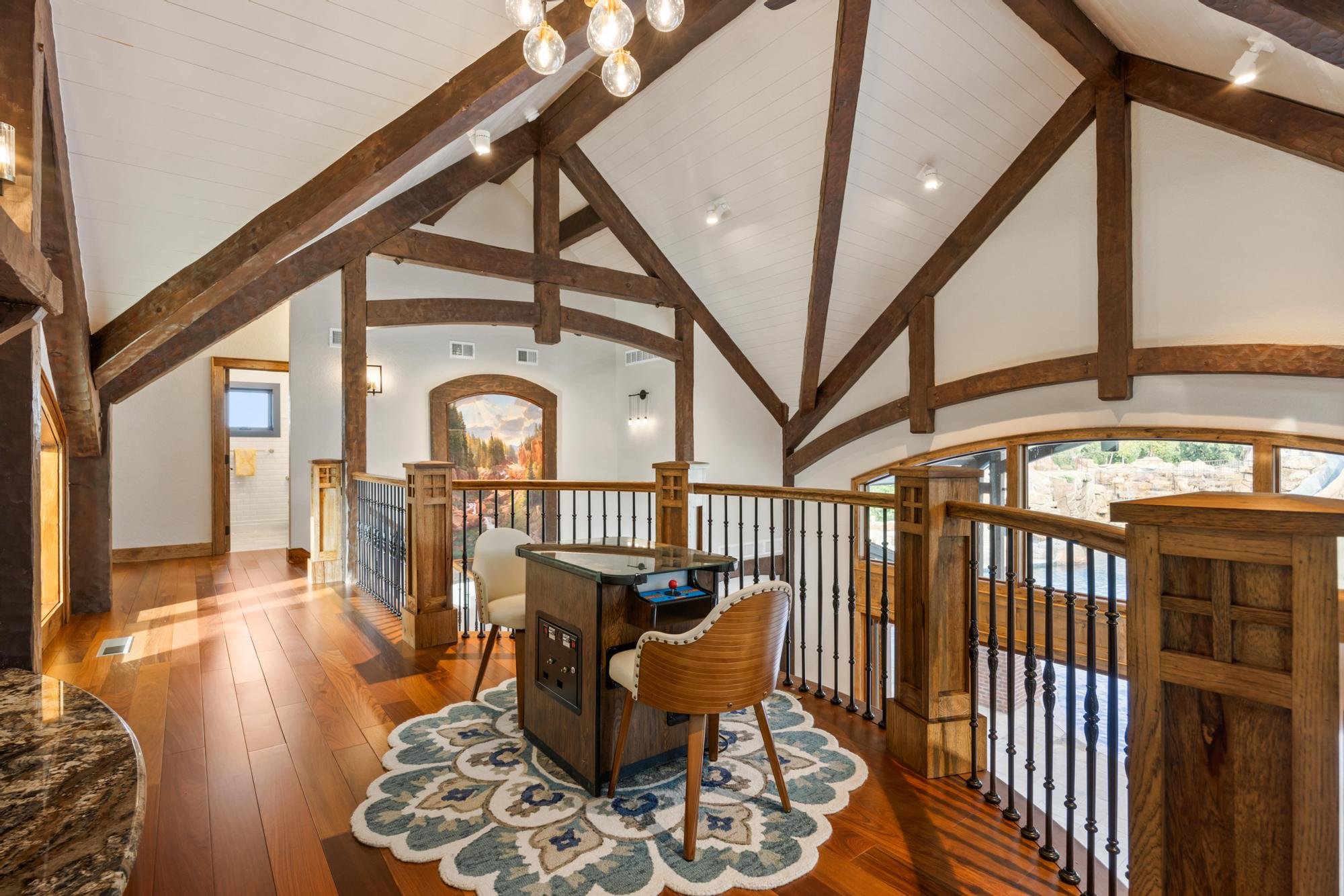 Interior of a spacious house with vaulted ceiling, exposed wooden beams, hardwood floor, and large windows showing a scenic view. There is a sitting area with two chairs, a small table, and a patterned rug.