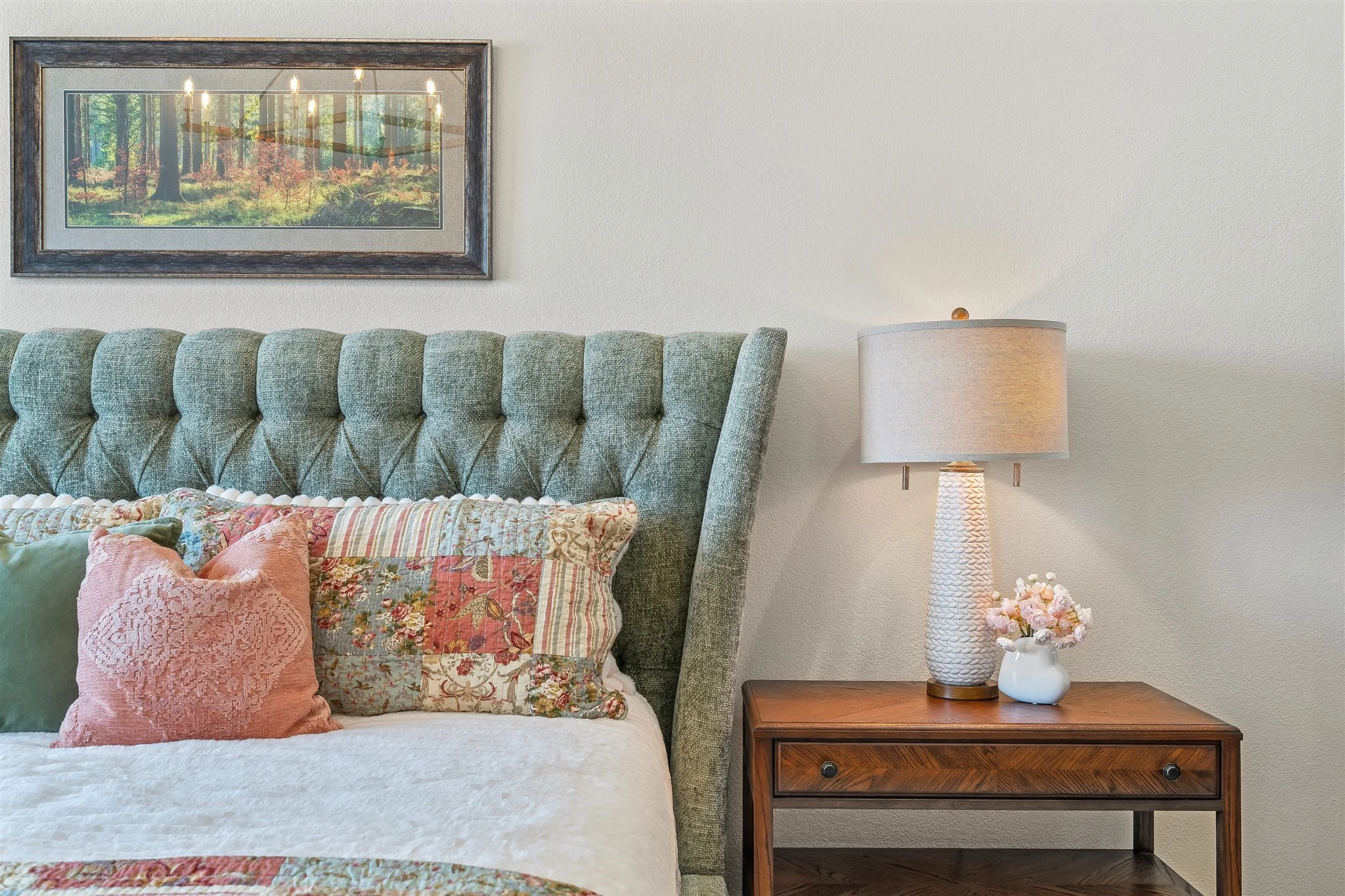 A bedroom with a green upholstered headboard, decorated pillows, a lamp on a wooden nightstand, a framed forest landscape photograph on the wall, and a small vase with pink flowers on the nightstand.