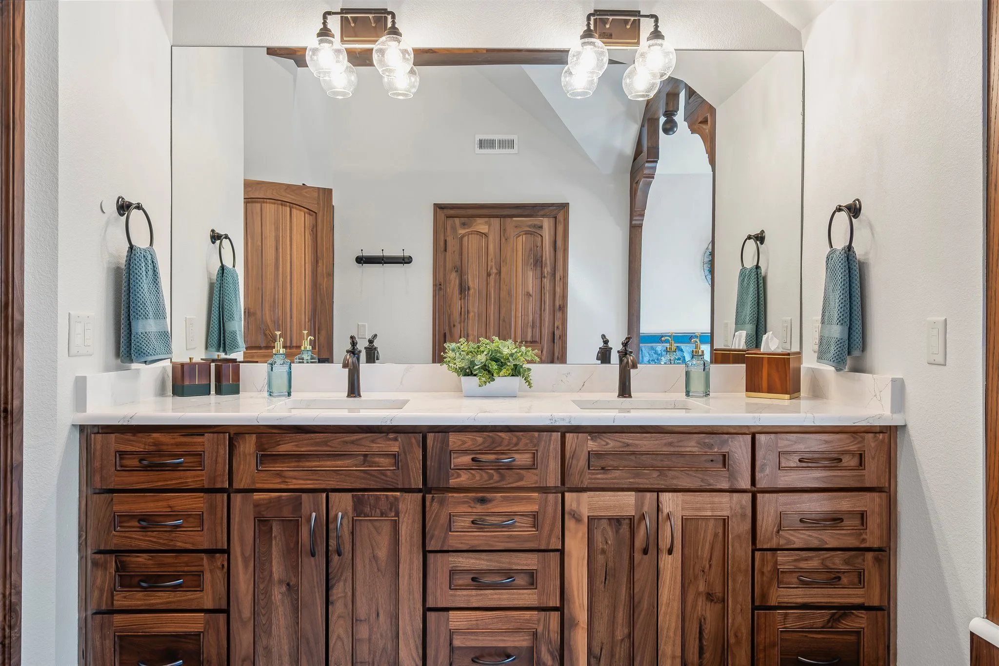 Bathroom vanity with wooden cabinet, large mirror, two sinks with black fixtures, decorative plants, soap dispensers, and blue towel hanging on wall.