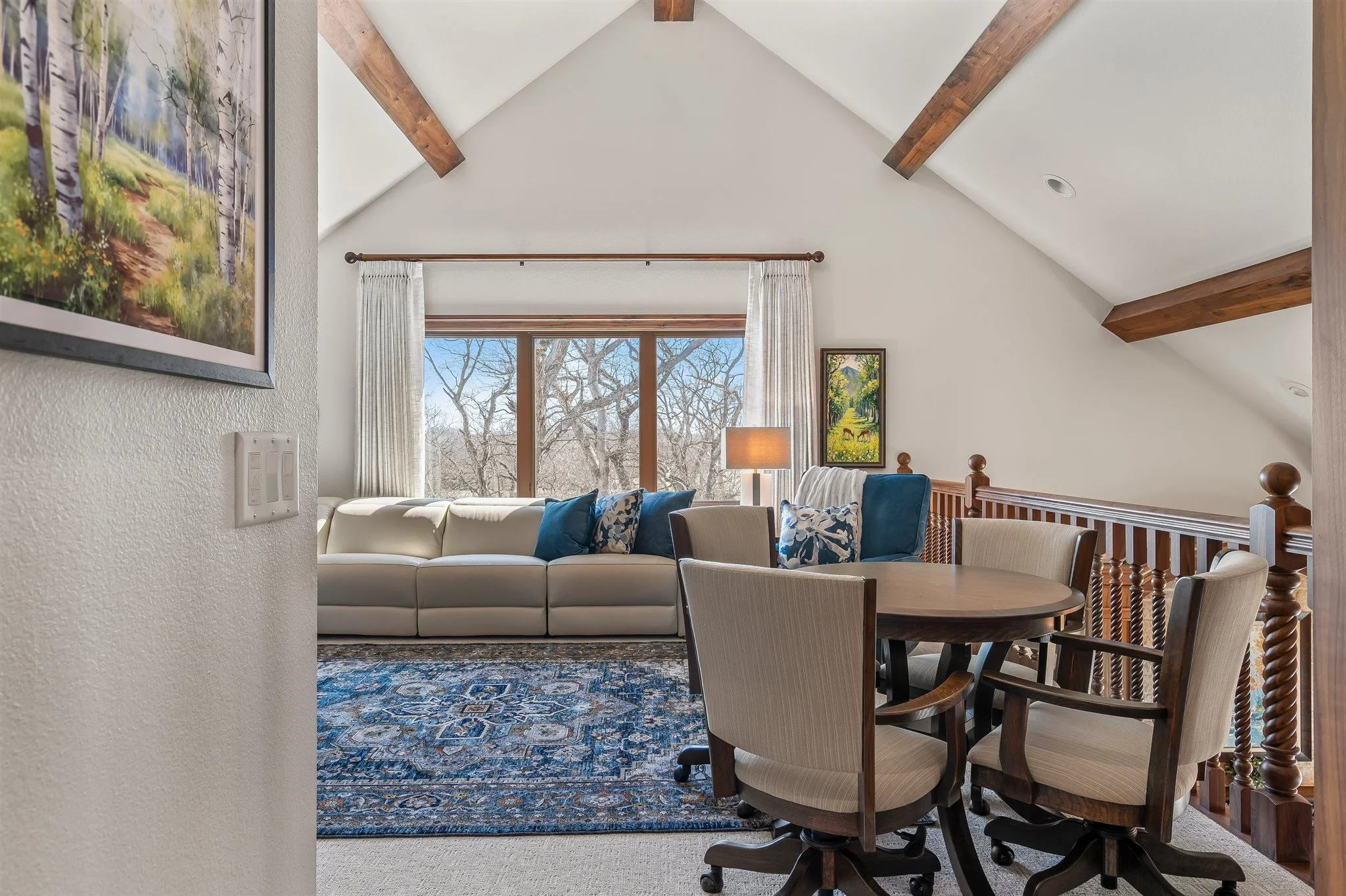 Living room with white sofa, blue throw pillows, and wooden dining table with beige chairs near a railing. Large window with curtains showing snow-covered trees outside, artwork on the walls, ceiling beams, and a patterned rug.