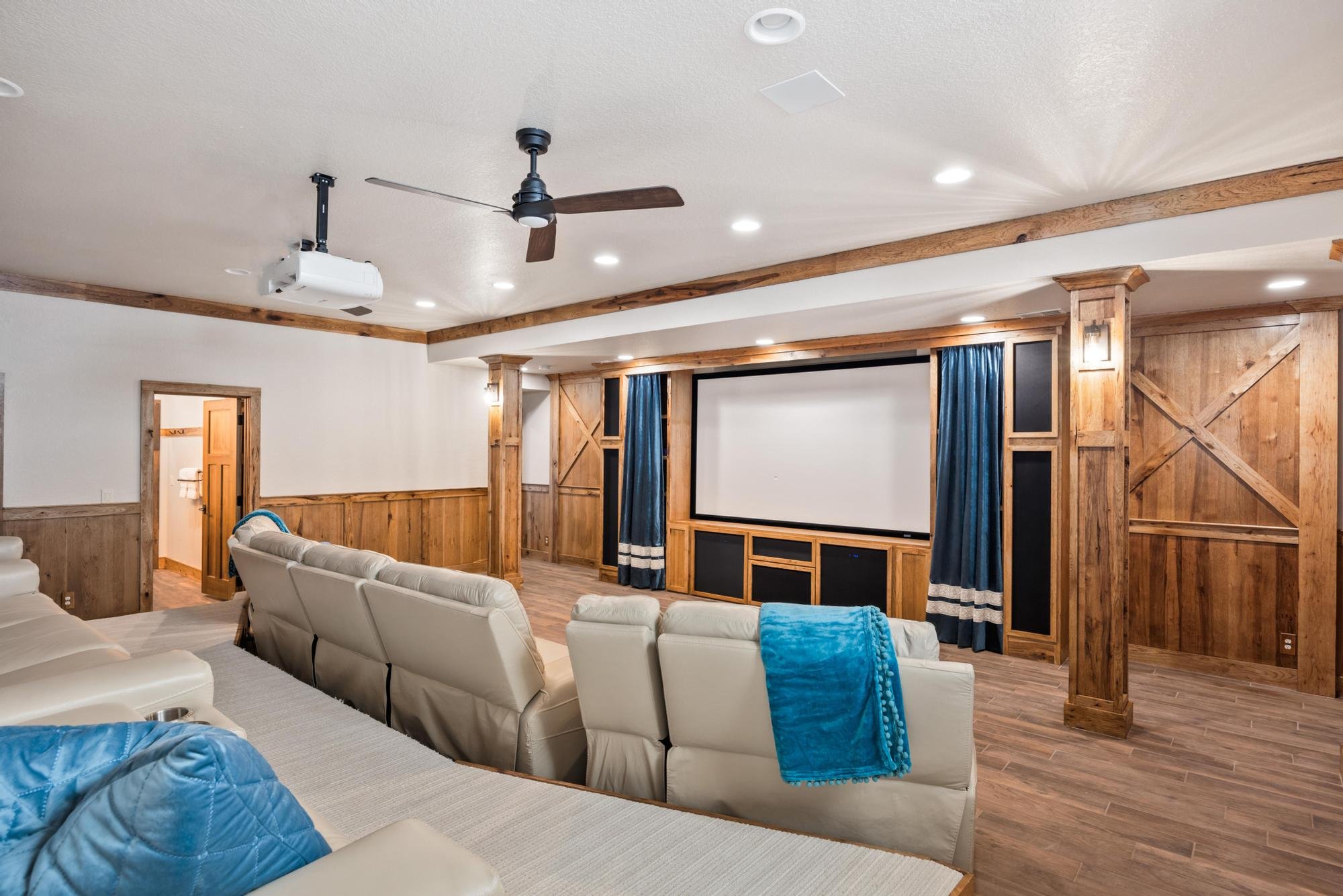 Living room with white leather recliners, a large wall-mounted screen, wooden paneling, and blue curtains, with a ceiling fan and projector.