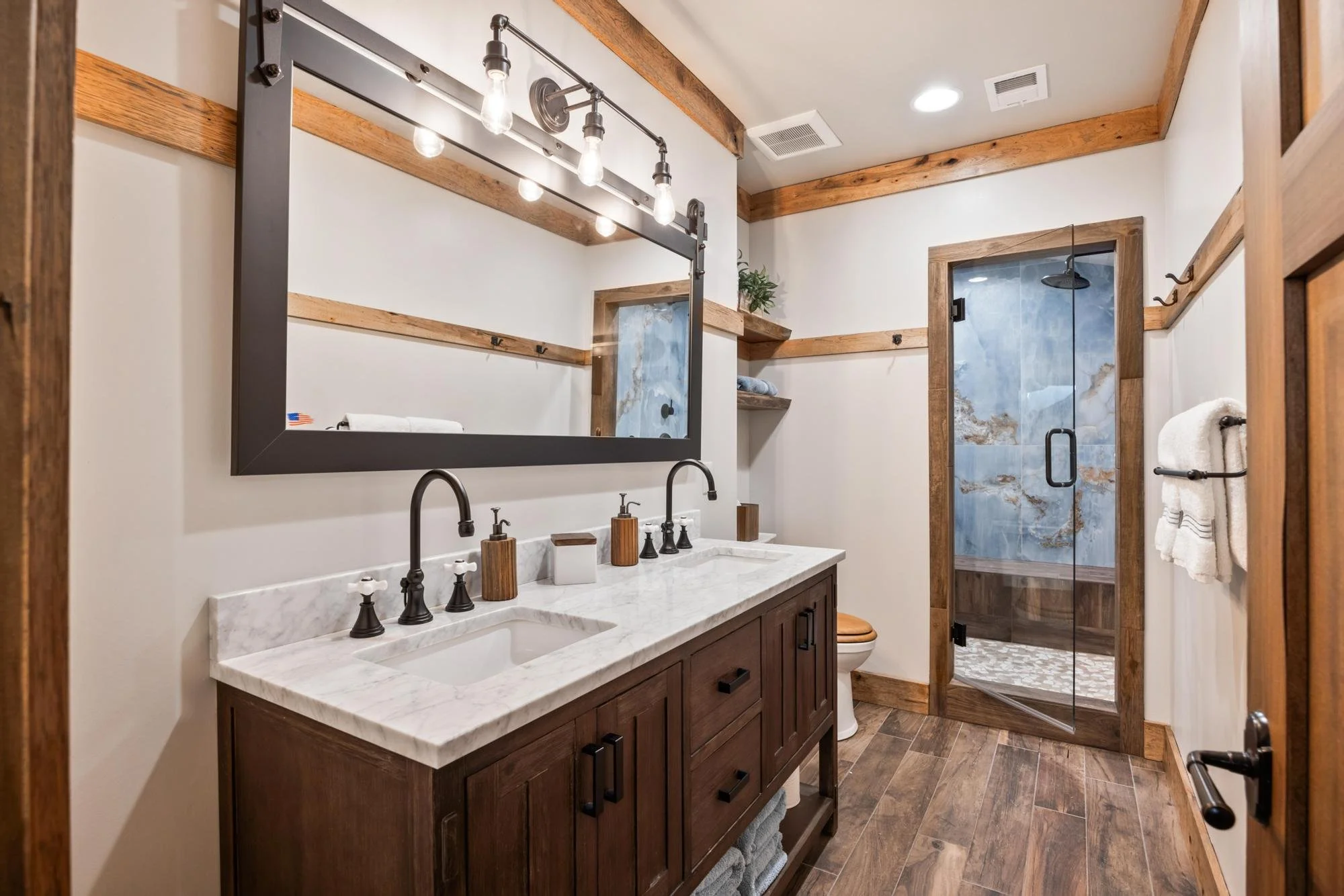 The image depicts a rustic bathroom with a dual-sink vanity topped with a white marble countertop, dark wooden cabinet, and black faucets. Above the sinks is a large rectangular mirror framed in black, with industrial-style light bulbs hanging above.