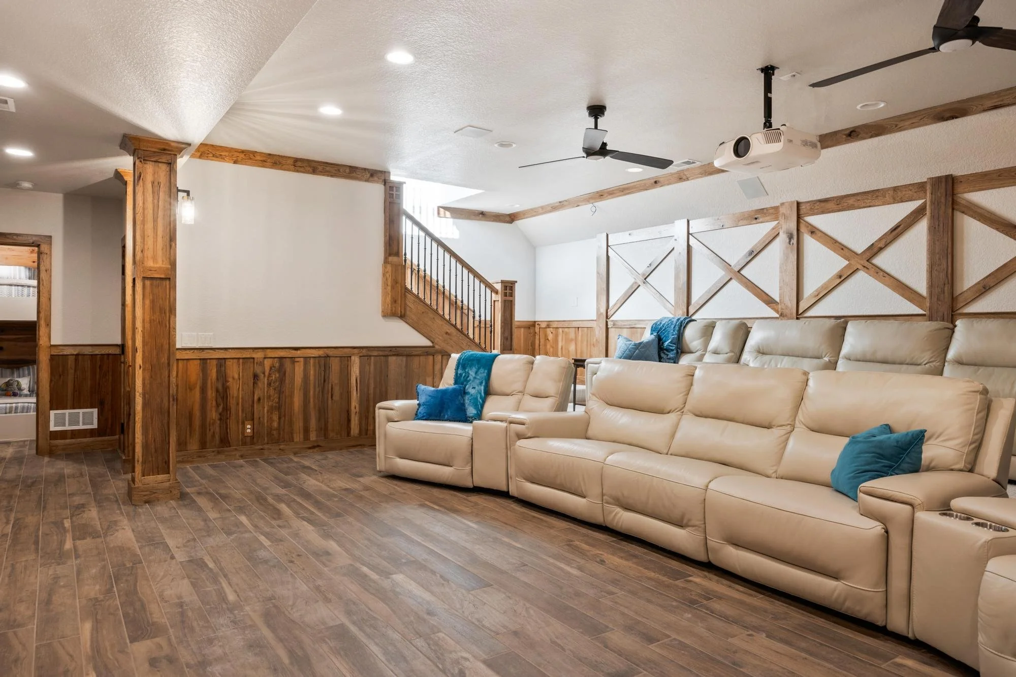 Living room with beige leather sofa with blue pillows, wooden wall paneling, ceiling fans, and a mounted projector.