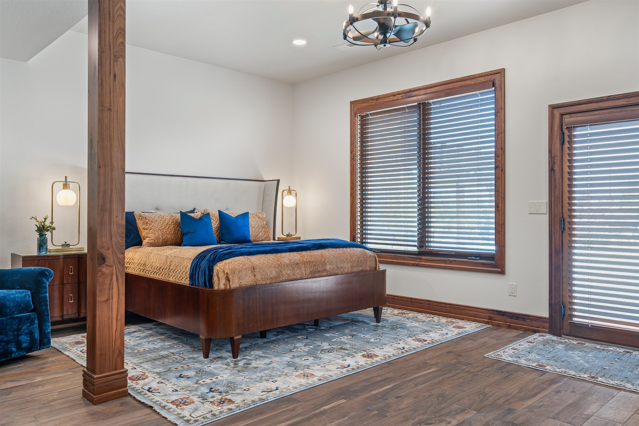 A bedroom with a wooden bed frame, tan bedding, and blue pillows, flanked by two wall-mounted lamps. Large windows with wooden frames and blinds, a patterned area rug, and a wooden post near the bed, with a modern chandelier overhead.