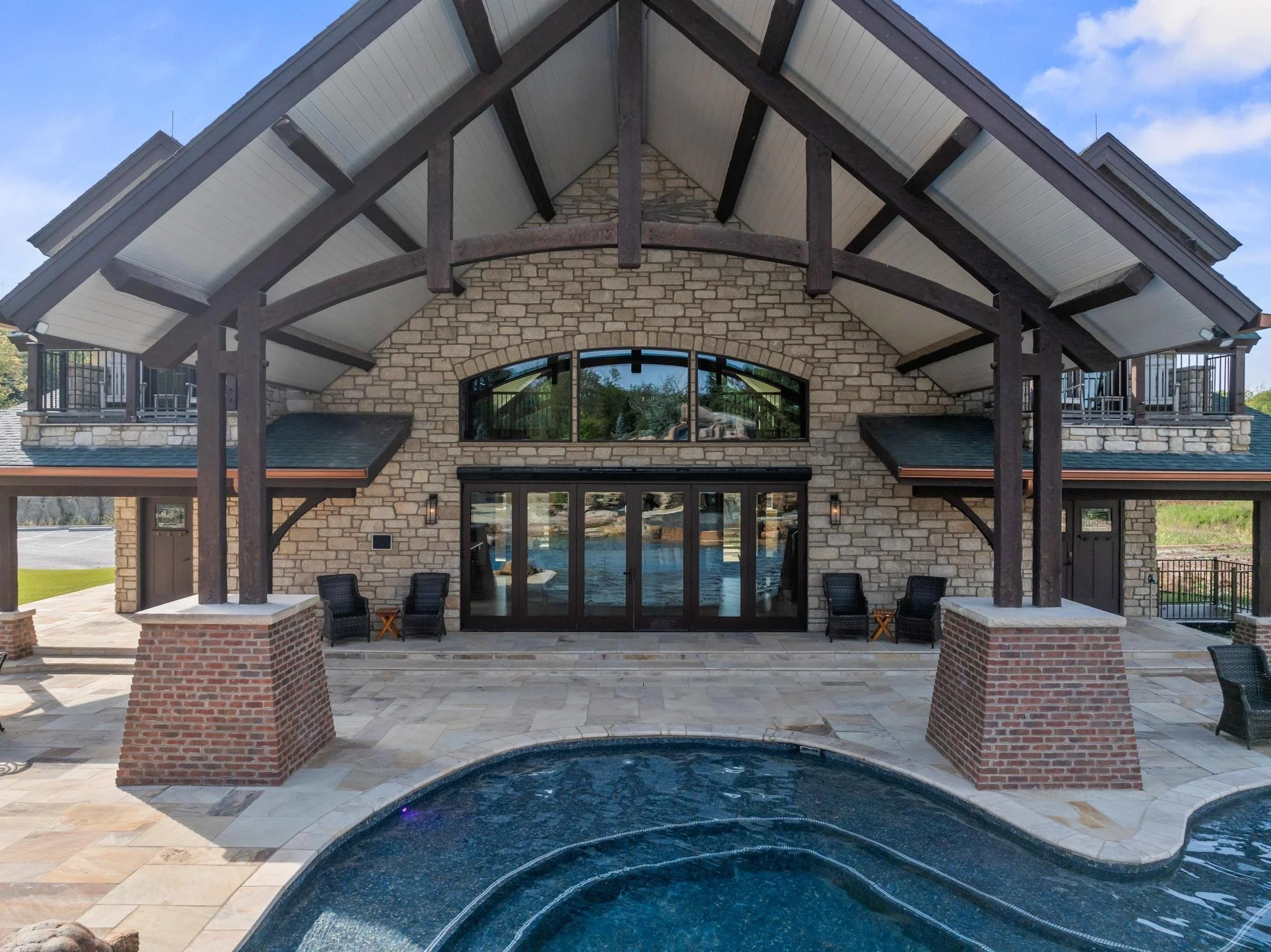 A house with a brick and stone exterior featuring a large arched window and a wooden pergola with a gable roof, overlooking a water feature and patio seating area.