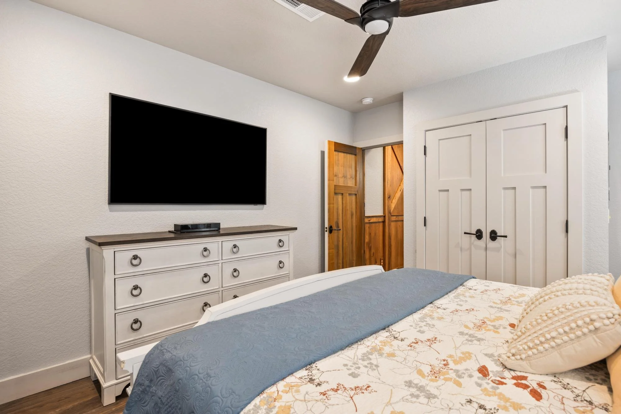 A bedroom with a bed covered in a bedspread with floral design and a blue blanket, a white dresser underneath a flat-screen TV, wooden doors, and a ceiling fan.