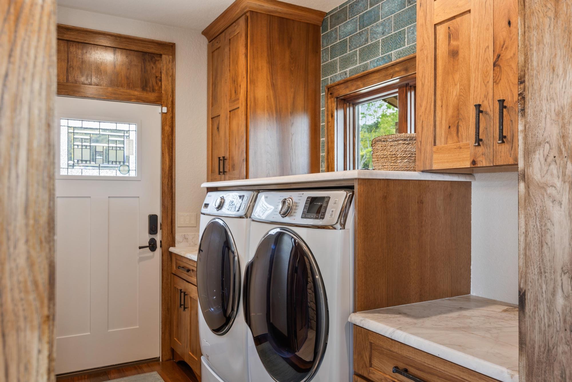 A laundry room with wooden cabinets, a washer and dryer, a small window showing trees outside, and granite countertops.