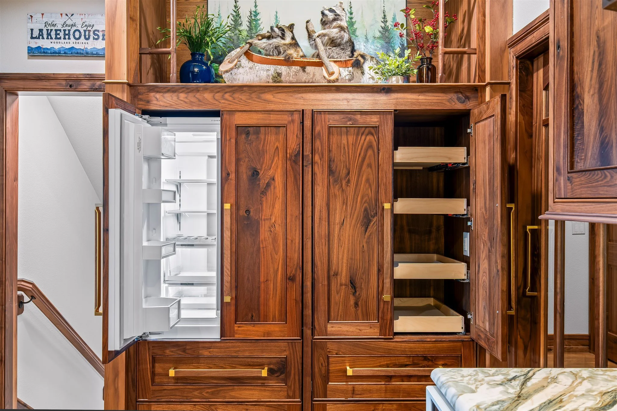 A wooden cabinet with two open drawers, a built-in refrigerator, and additional empty shelving space. There is a wallpaper mural of a forest scene with two raccoons on a tree branch, positioned above the cabinet. To the left, a decorative poster with