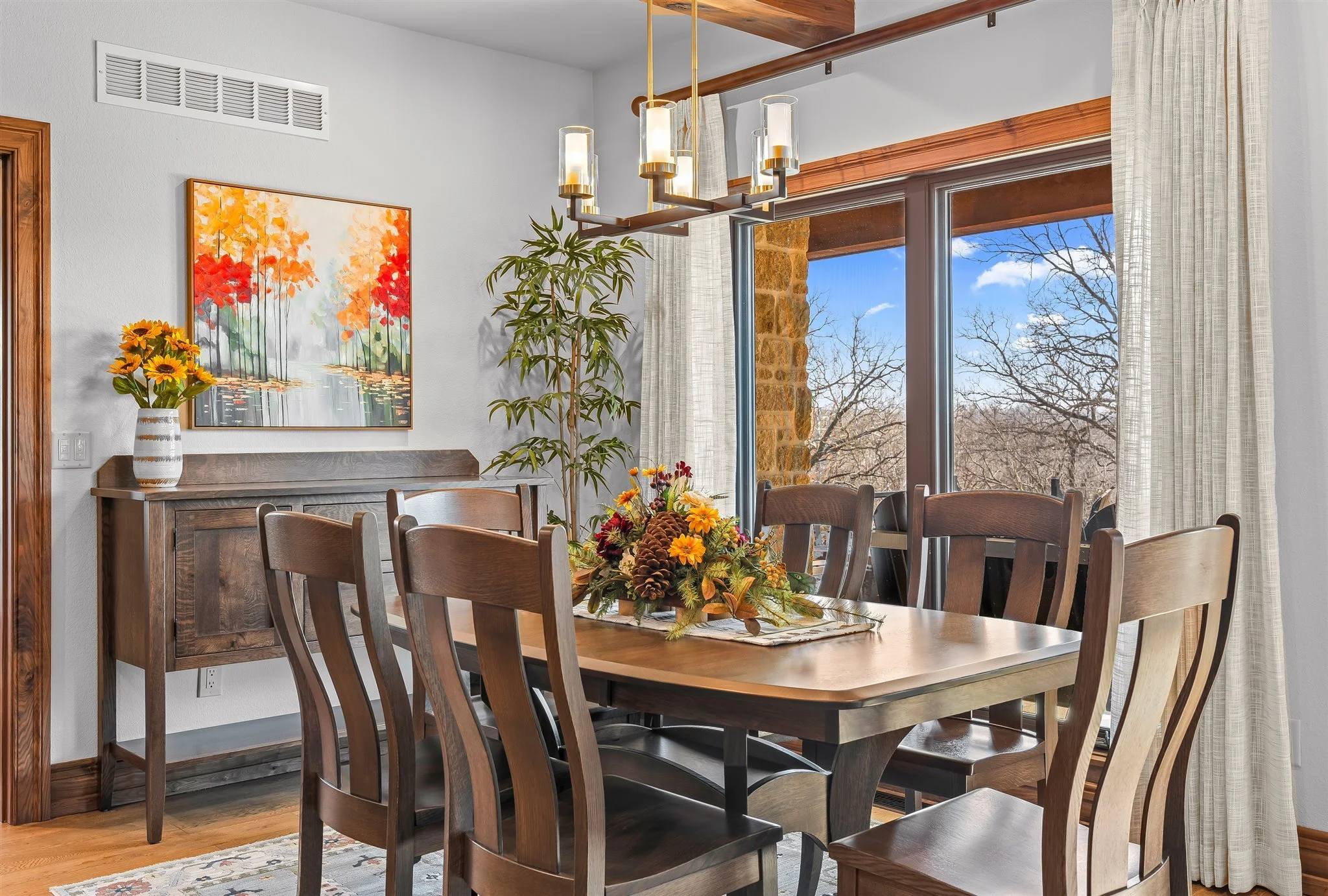 A wooden dining table with a floral arrangement in autumn colors, surrounded by wooden chairs, in a dining room with large windows showing a clear blue sky and leafless trees outside. There is a hanging chandelier above the table, a painting of color