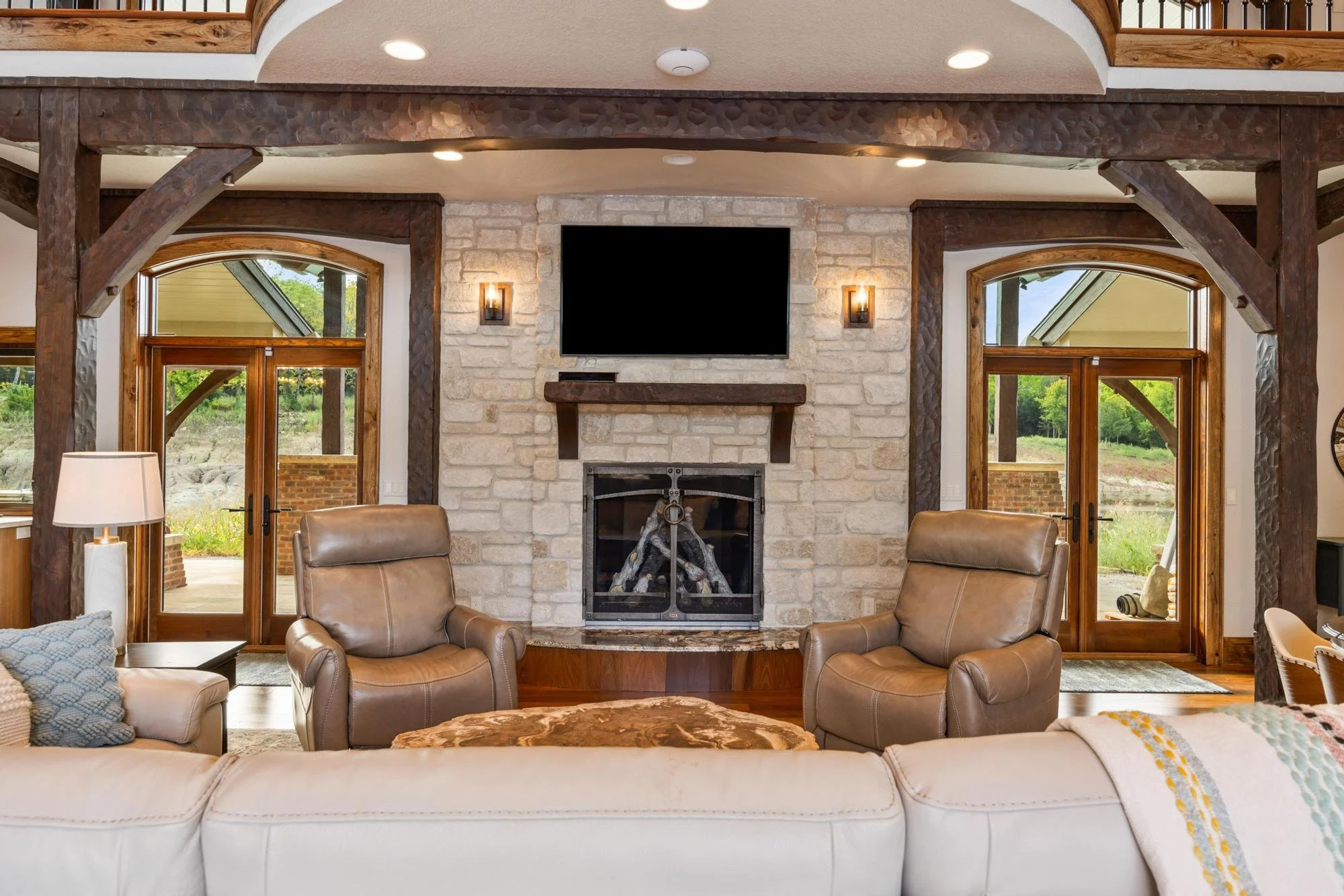 Living room with two leather armchairs around a stone fireplace, mounted flat-screen TV, large windows, wooden beams, and a porch outside.
