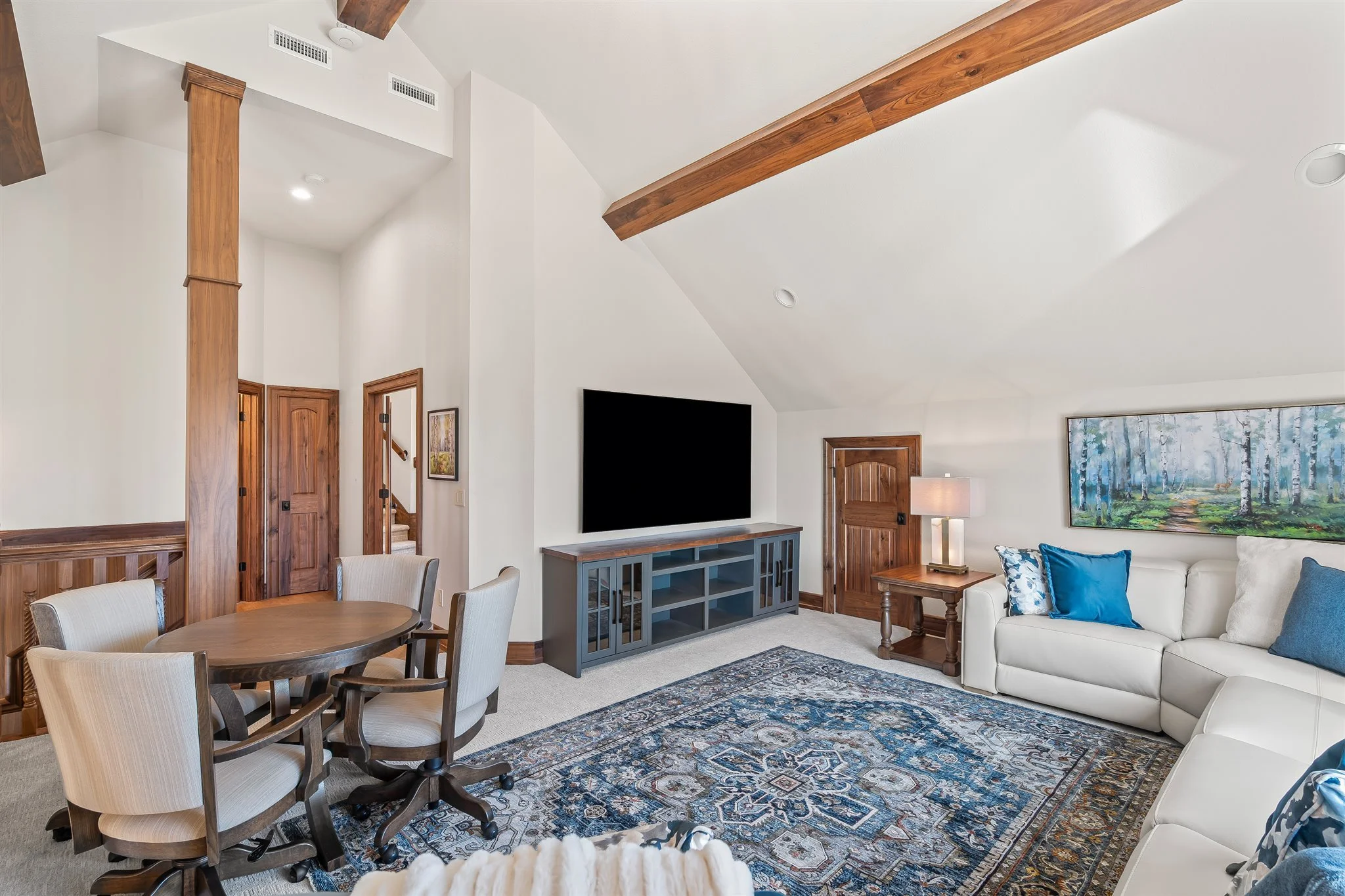 Living room with a white sofa, decorative pillows, a wooden side table with a lamp, a painting of a forest, a television on a blue stand, a round dining table with beige upholstered chairs, wood accents on doors and beams, and a multicolored area rug