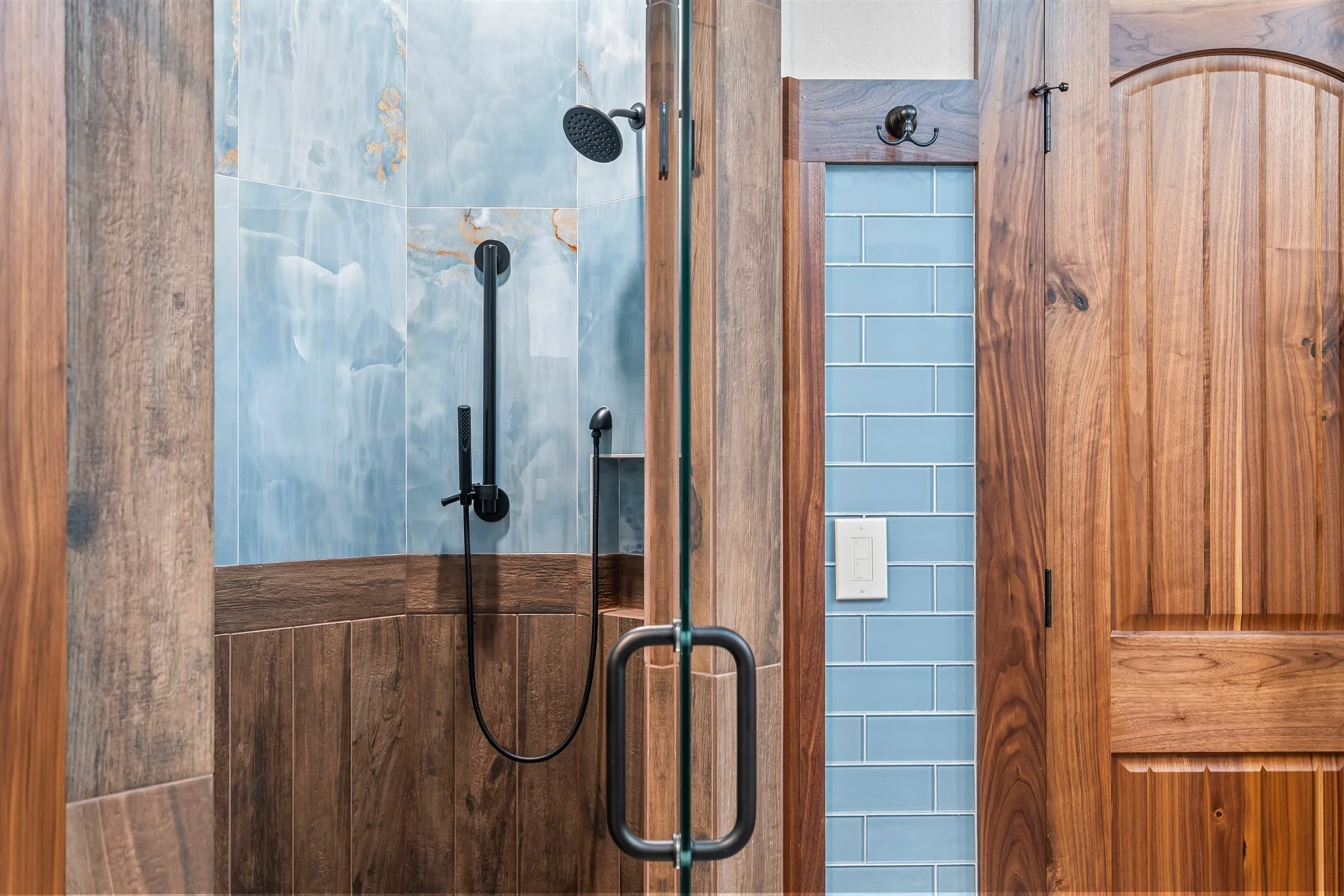 Interior view of a bathroom shower with light blue tiles, a black showerhead, a handheld shower wand, and a wooden frame. Part of a wooden door with a black handle is visible on the right side.