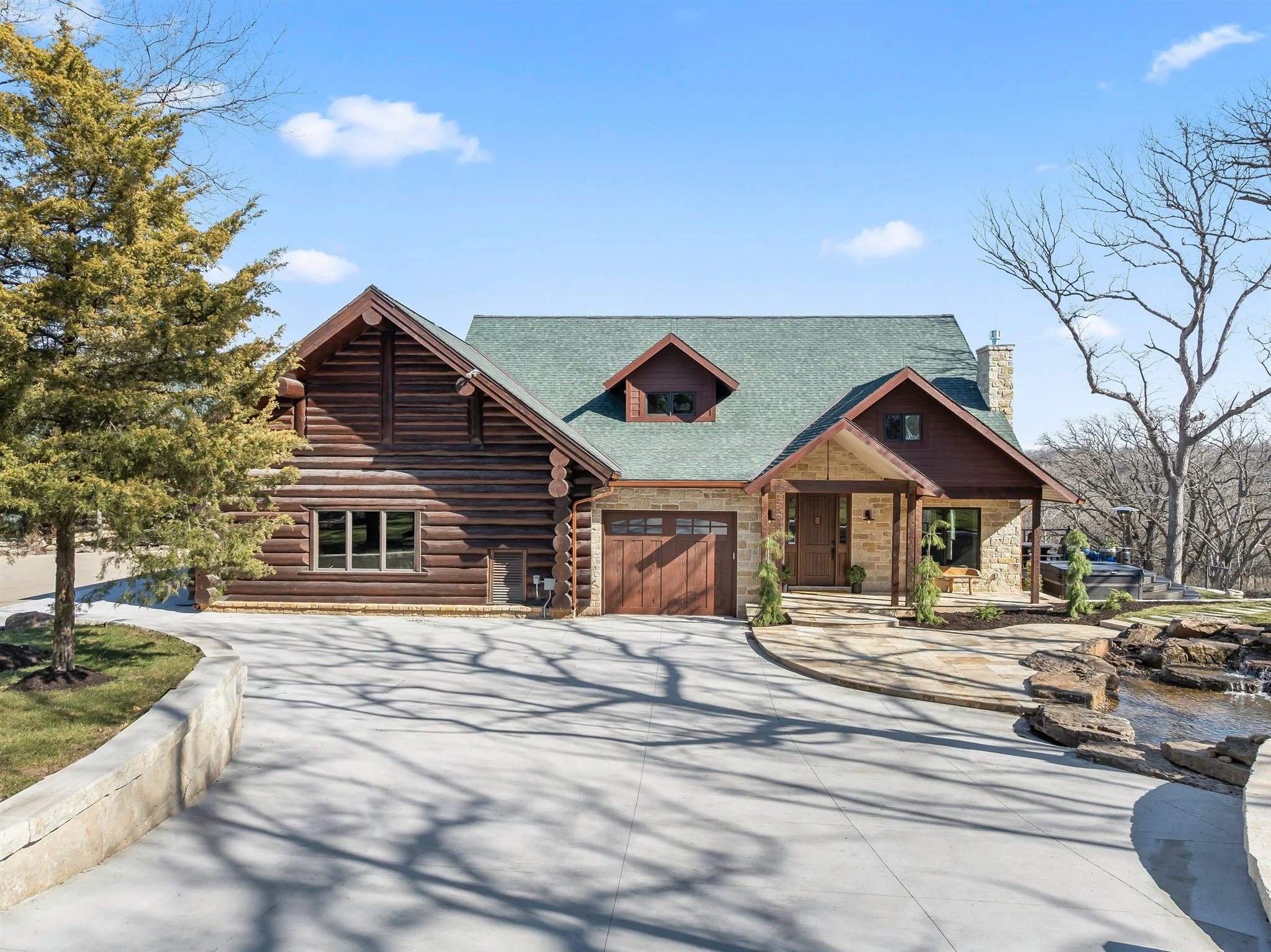 A house with a green roof, brick and wood exterior, and a garage door, surrounded by trees and a paved driveway under a blue sky with clouds.