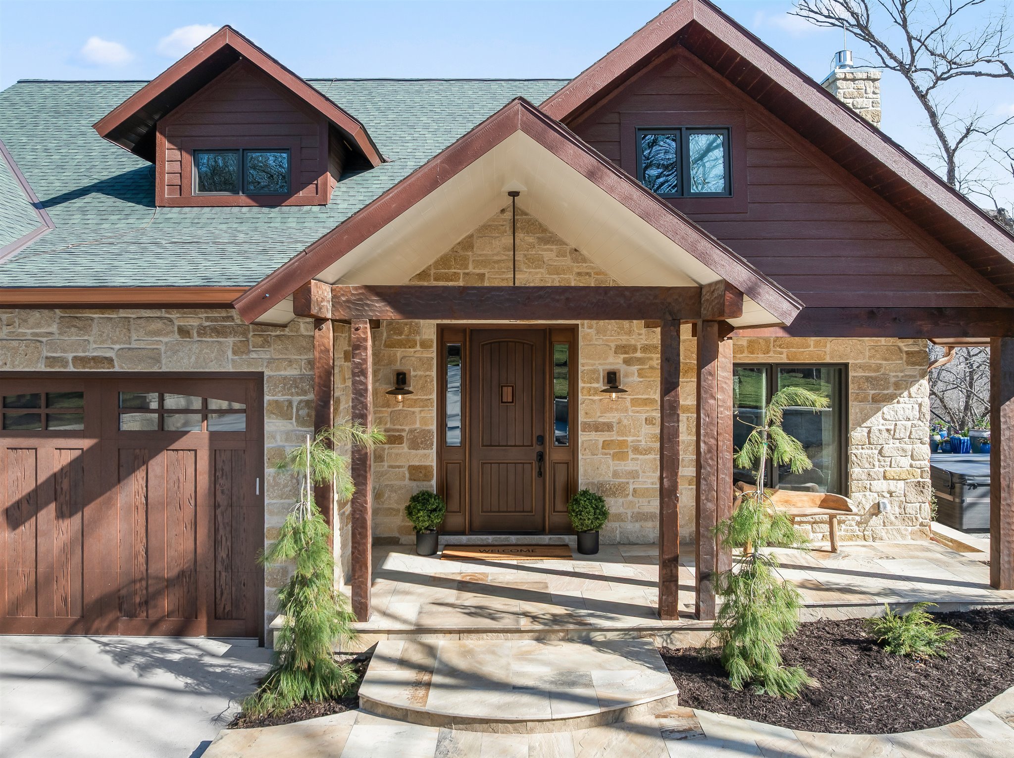 Front entrance of a house with a stone and wood exterior, a wooden door with a 'Welcome' mat, and a covered porch supported by wooden beams. There are two potted plants at the entrance, and a paved walkway leading to the door. The house has a gabled 