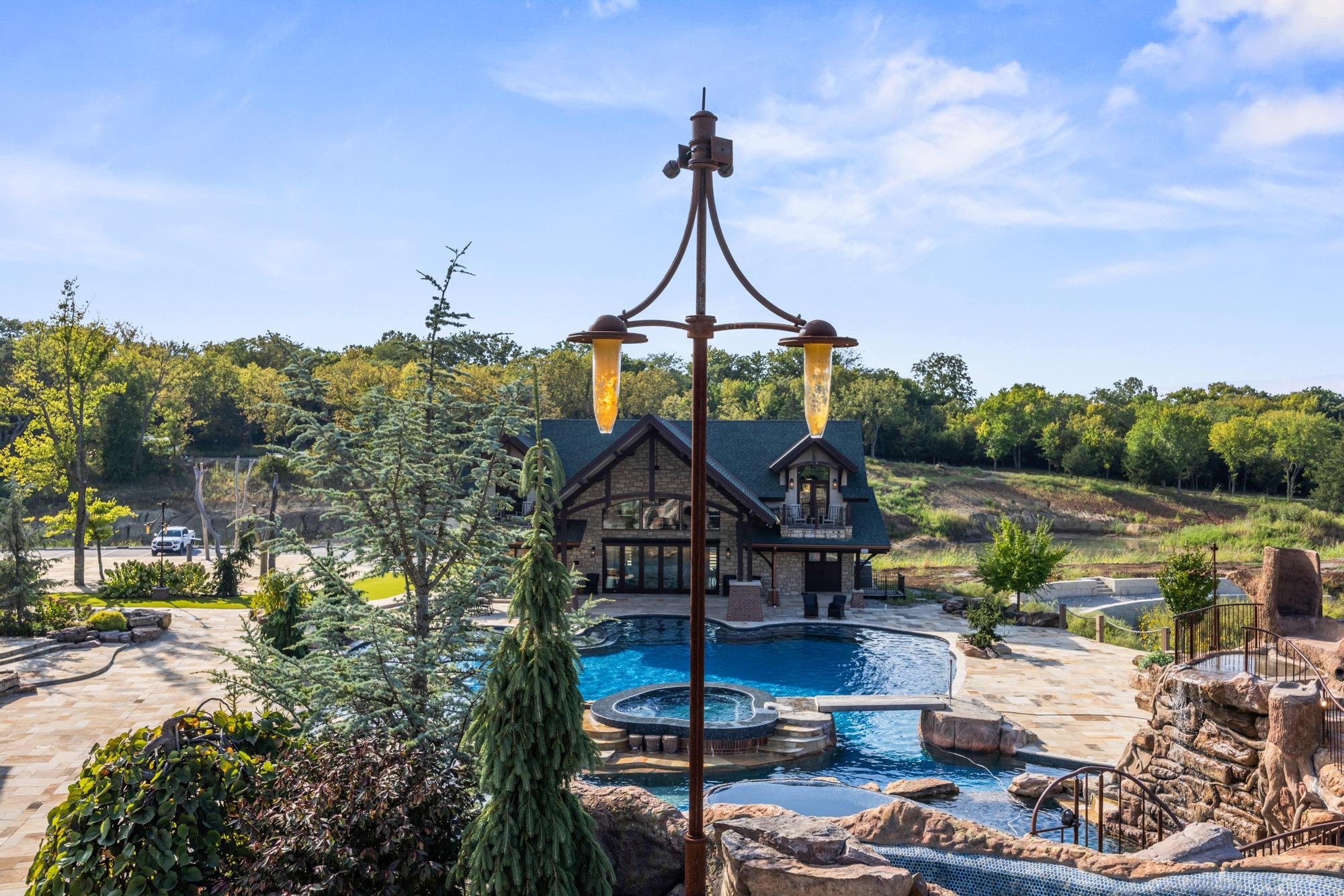 A backyard with a swimming pool, hot tub, stone patio, and a house in the background surrounded by trees and greenery on a sunny day.