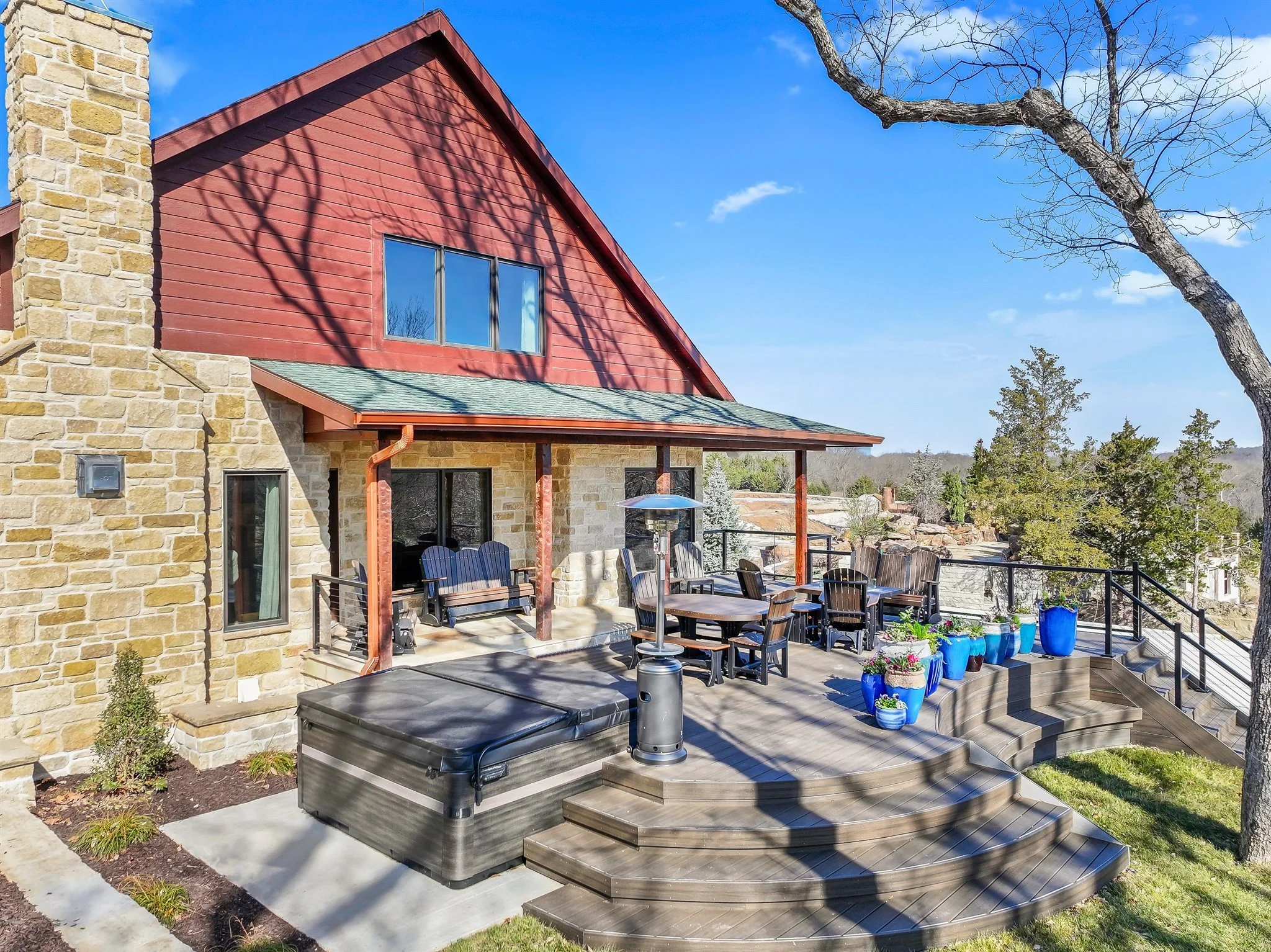 A backyard patio with wooden decking, blue flower pots, outdoor dining table and chairs, a tall heater, and a stone house with red siding on the upper level. There is a bare tree casting shadows on the house, and a clear blue sky in the background.