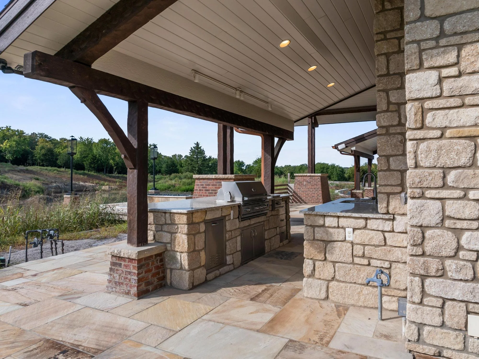 Outdoor kitchen with stone and brick construction, built-in grill, and covered patio with recessed lighting, overlooking a grassy landscape.