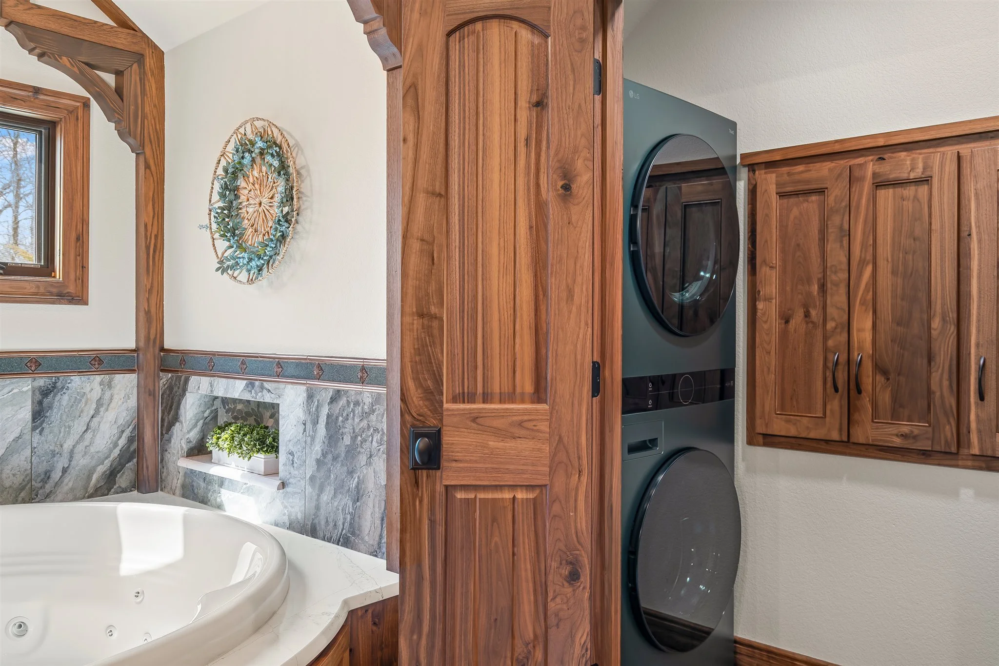 A laundry area with a stacked washer and dryer set, wooden cabinets, and a wall with a beige textured surface. There is a decorative wreath on the wall and a window with natural light.