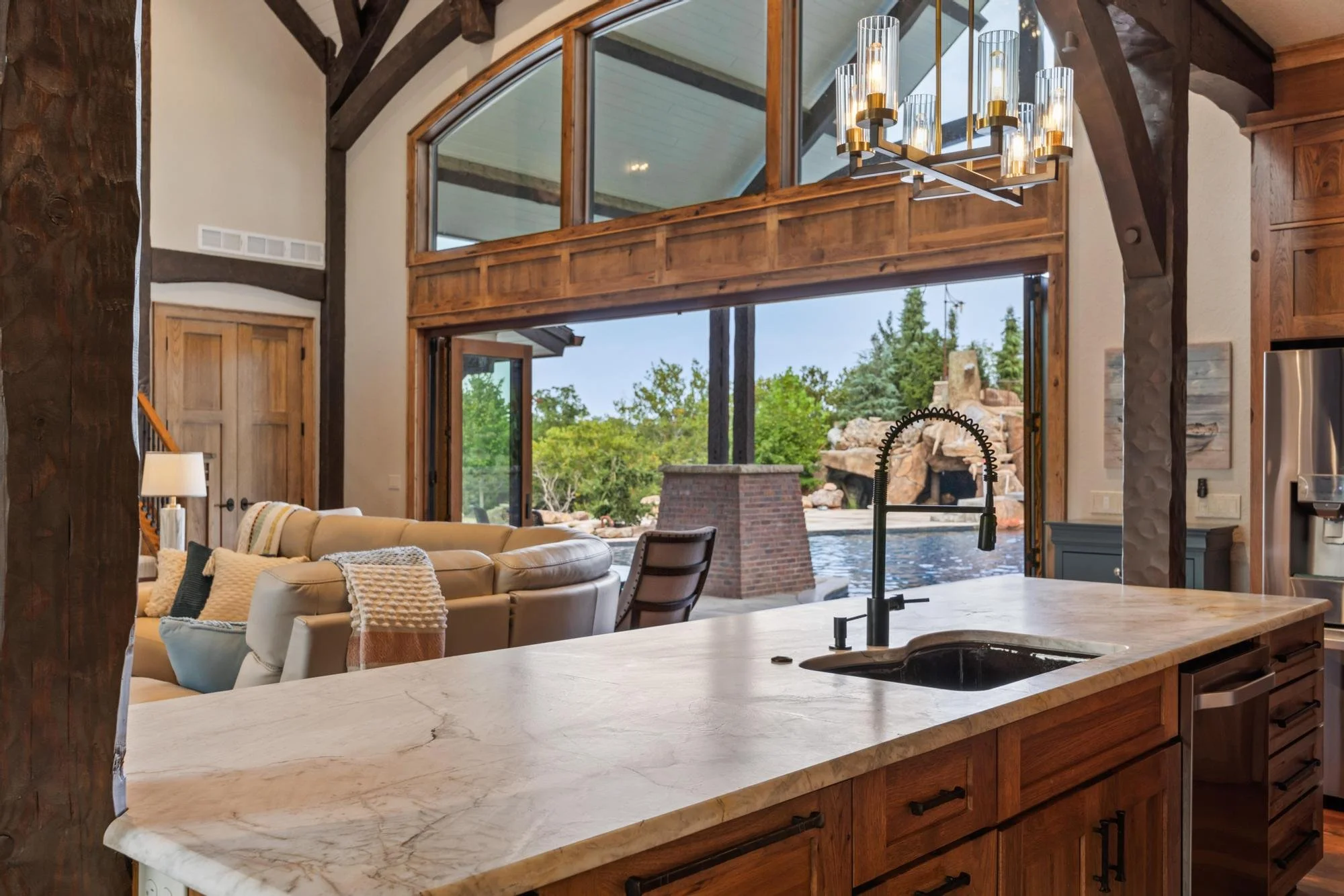Open-concept living area with large windows showing outdoor trees and rocks. Kitchen island with a marble countertop, black faucet, and kitchen appliances in the background. Cozy seating with pillows and a lamp.