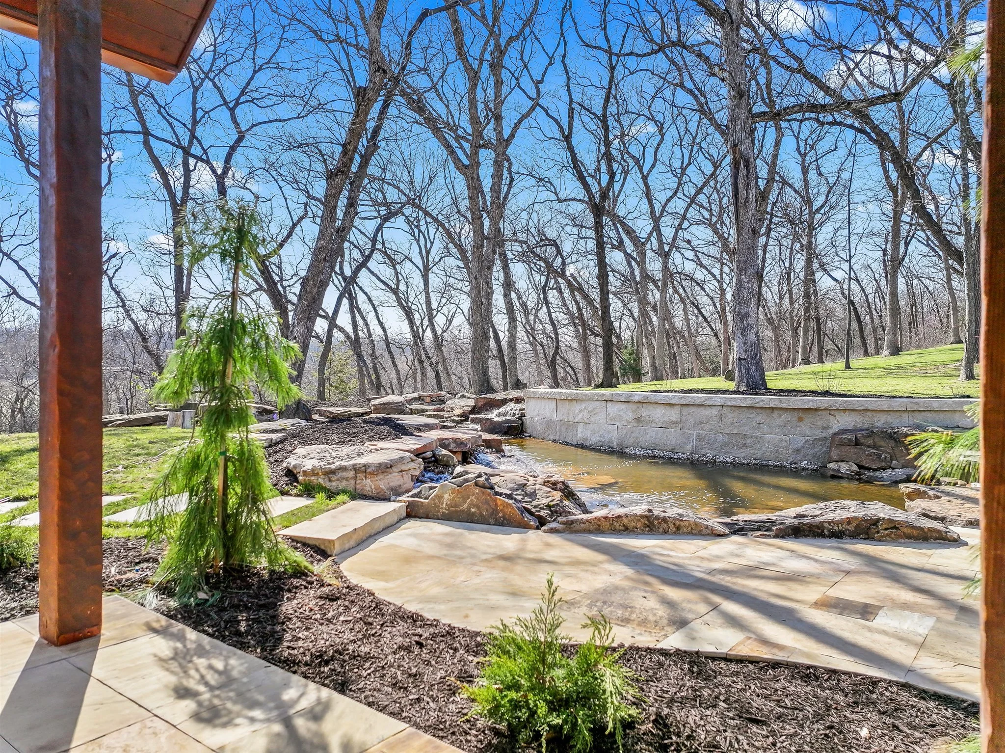 View of trees and a small stream, seen from a covered porch with a wooden post and part of a roof in the foreground. Clear blue sky in the background.