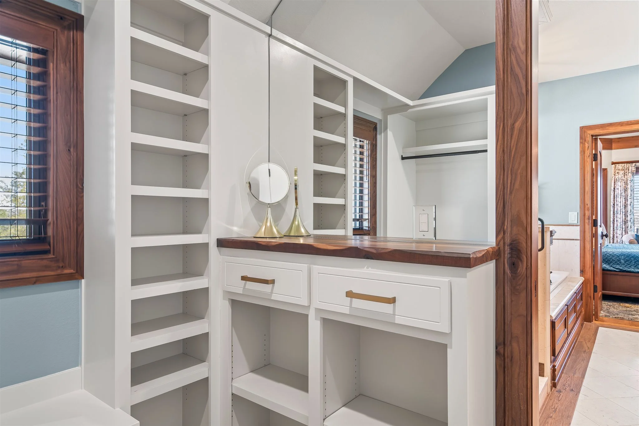 Empty built-in white cabinets with open shelves and drawers, situated near a window with wooden blinds, and a partial view of a bedroom with floral curtains.