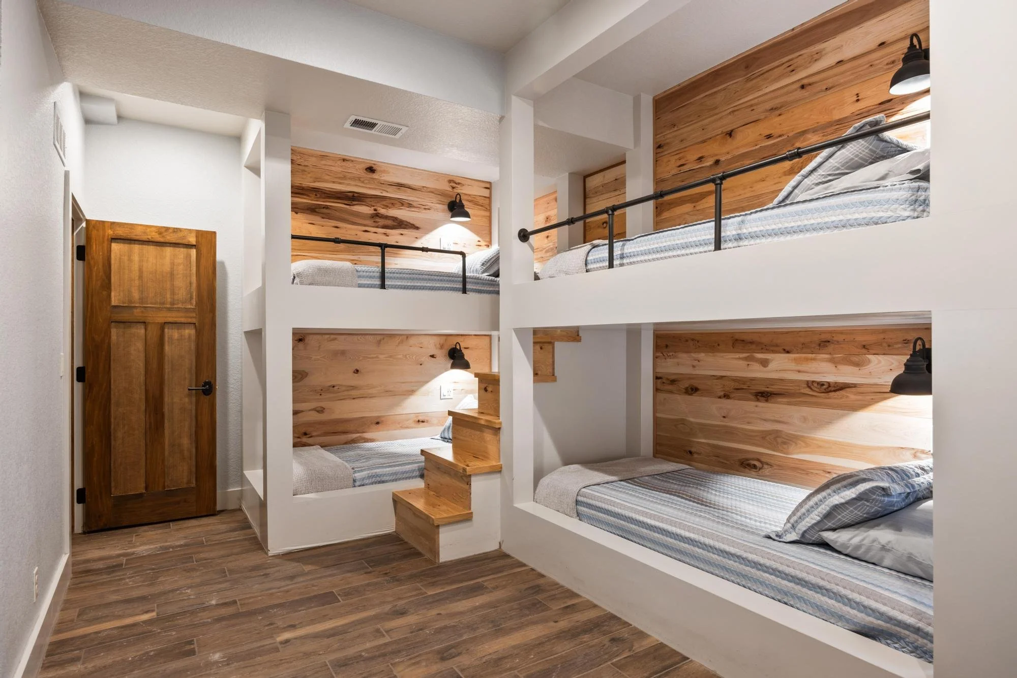 Photo of a bunk bed with four sleeping spaces, featuring wooden headboards and striped bedding, in a bedroom with wood wall paneling and black wall-mounted reading lamps.