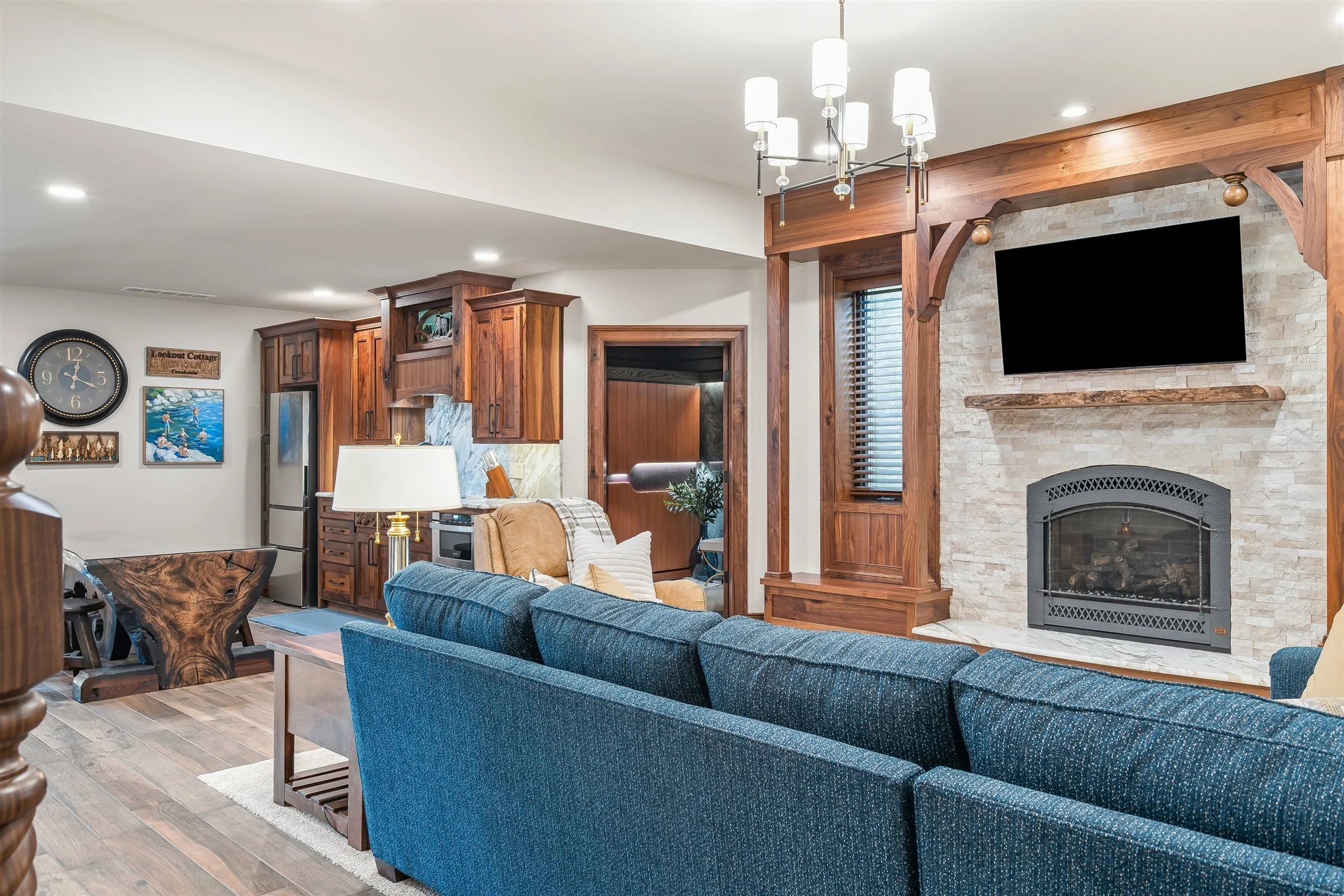 Interior view of a living room with a stone fireplace, a flat-screen TV, wooden trim, blue couches, a wooden coffee table, and various wall decorations, including a clock and paintings.