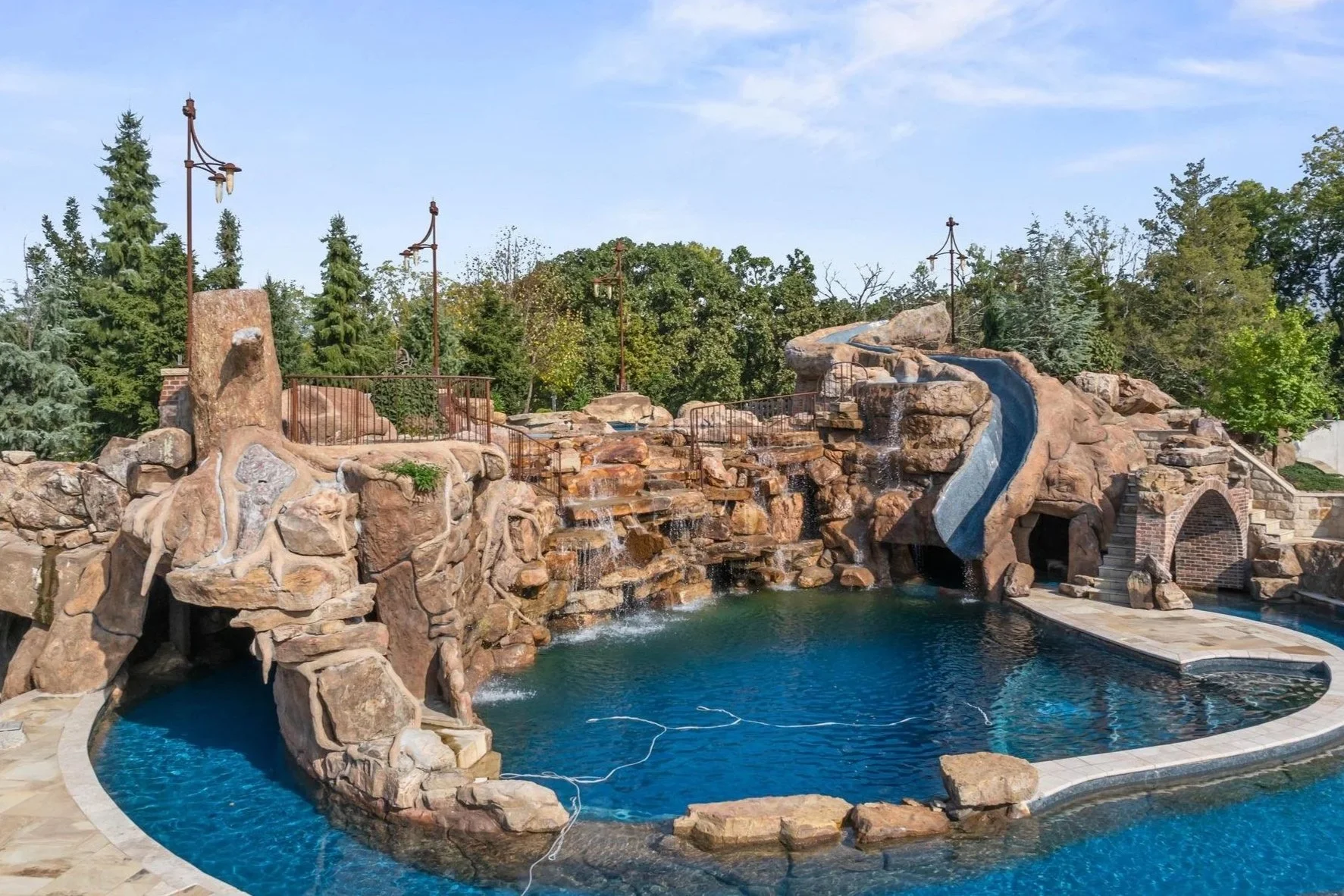 Empty backyard pool with rock waterfall feature, surrounded by stone patio, with a small water slide and trees in the background.