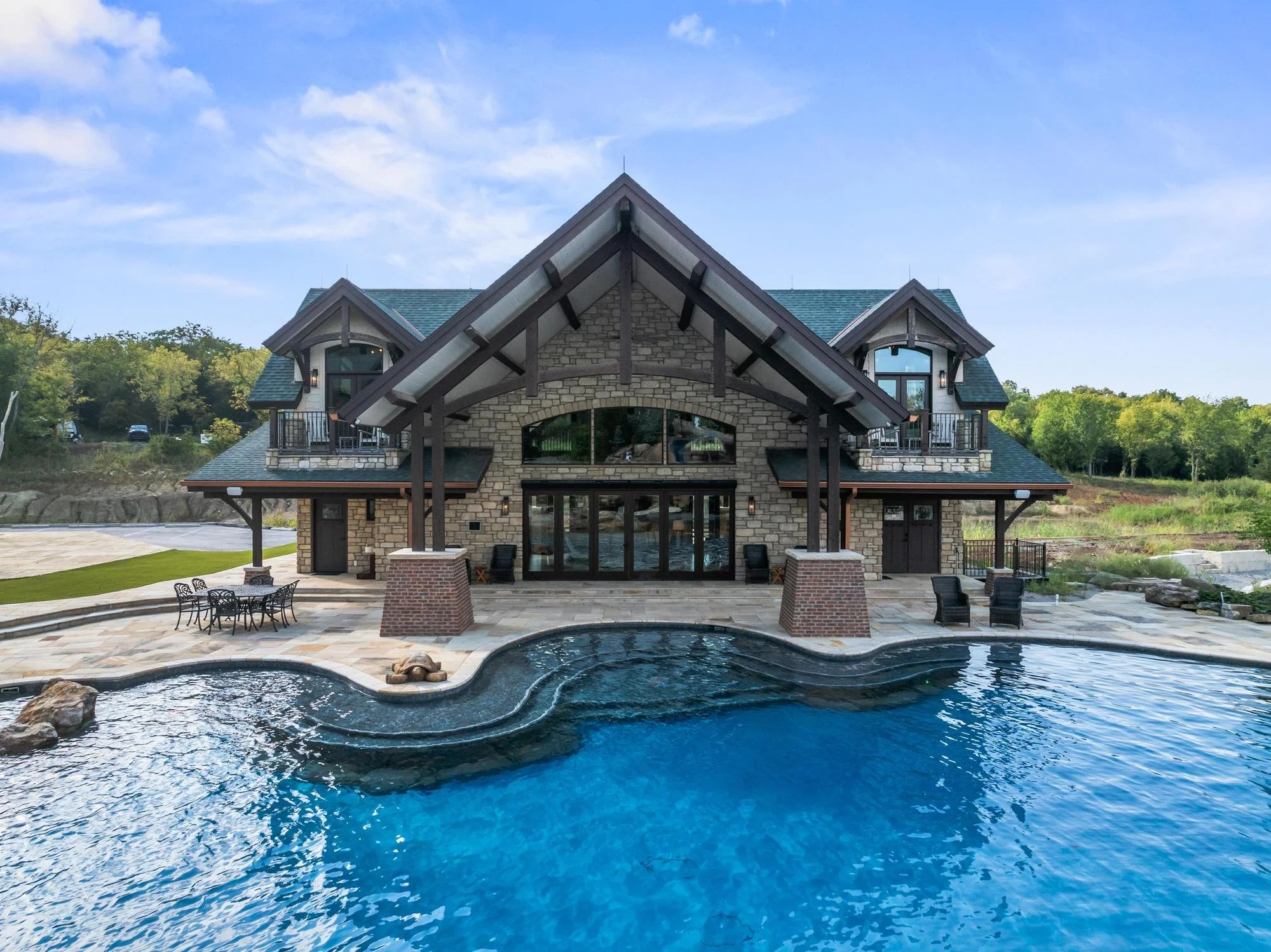 Large house with stone and wood exterior, multiple balconies, and a steeply pitched roof, situated near a landscaped pool with rock features and lounge chairs, surrounded by trees under a blue sky.