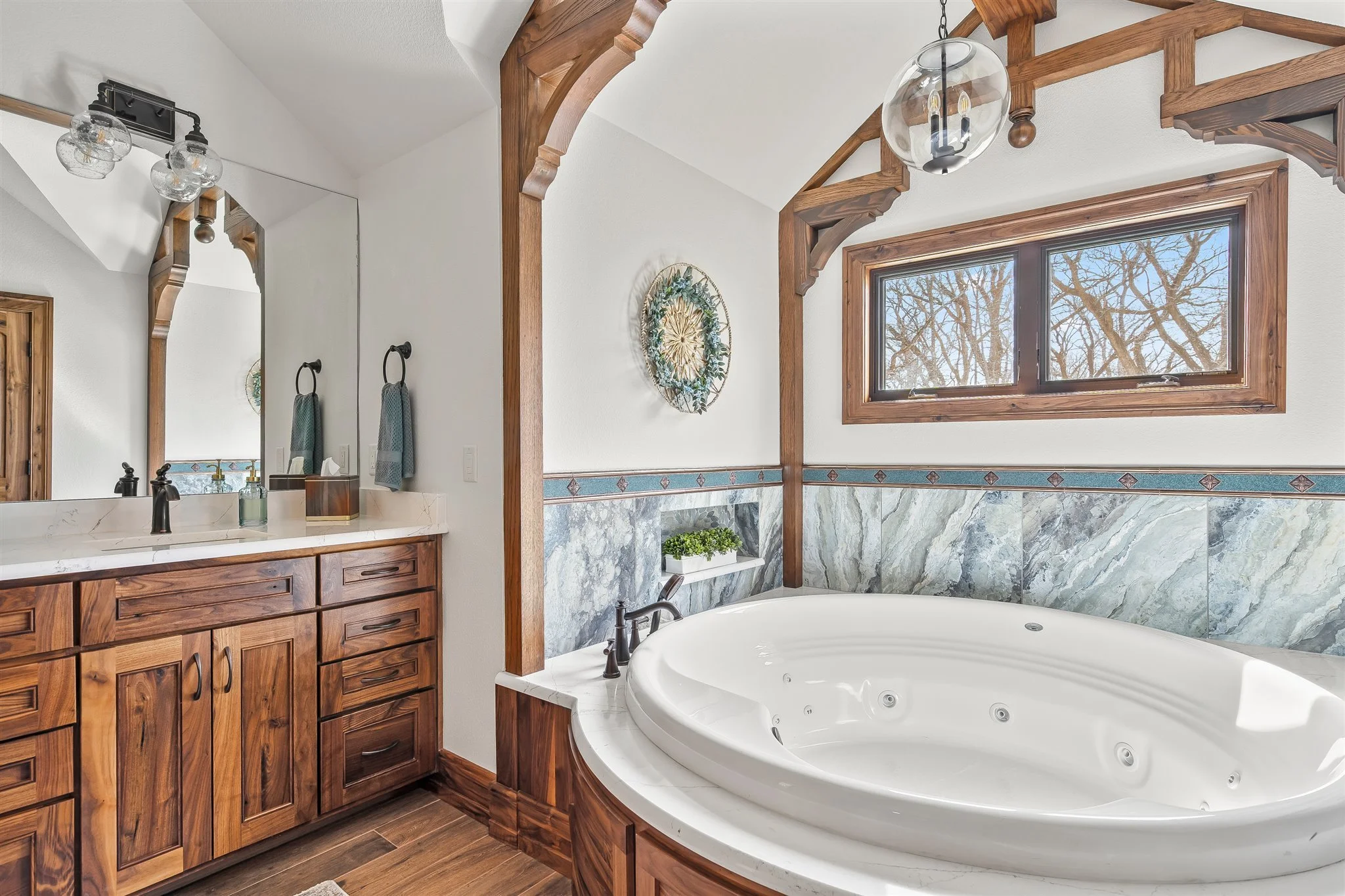 A bathroom with a large white soaking bathtub built into a corner, wooden cabinetry beneath a white countertop and a large mirror. There are two light fixtures above the mirror. A window next to the bathtub reveals tree branches outside. The wall fea