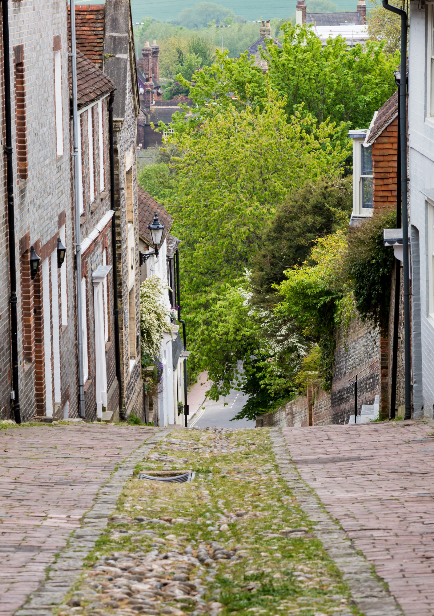 A cobblestone street descending downward between old brick and white painted houses with greenery and trees visible at the end of the street.